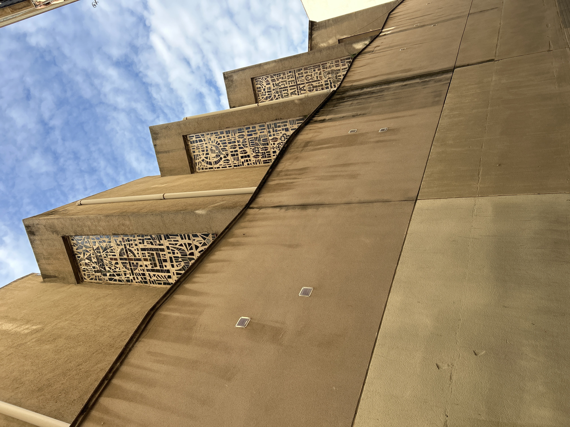 An upward-angled shot captures the distinctive stepped facade of a building against a bright blue sky with scattered white clouds. The building, likely made of concrete or stucco, is a warm sandy beige or light brown color, exhibiting a modernist or possibly brutalist architectural style.
The left side of the image features a series of staggered, recessed sections. Each of these sections contains a long, horizontal panel with an intricate, geometric pattern that resembles a decorative window or screen. These patterns are composed of various abstract shapes, including rectangles, arcs, circles, and irregular polygons, creating a complex tessellation in a light color against a darker background, possibly glass or void. A light-colored pipe runs horizontally along the lower part of one of these stepped sections.
A prominent diagonal line cuts through the image, separating the stepped, patterned side from a flatter, more uniform section of the building on the right. This flatter section also has a beige tone and shows some weathering and dark streaking, particularly in the upper parts. Several small, square, recessed light fixtures are visible on this plain wall section. The perspective emphasizes the height and architectural details of the structure. No human activity or visible text is present. The bright lighting suggests it is daytime. The scene evokes a sense of structured urban environment, typical of architecture that can be found in a city like Barcelona, Spain.