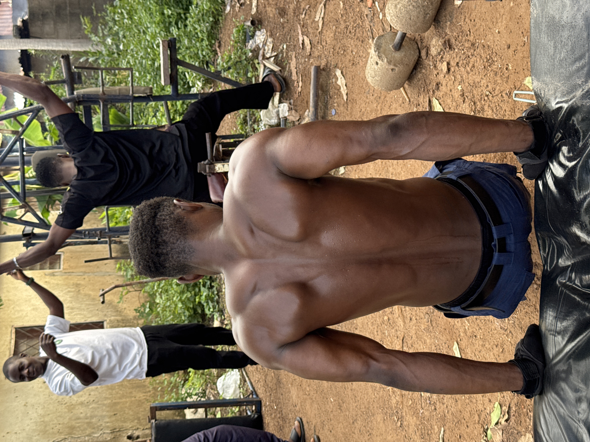 The image depicts three individuals in an outdoor, rural setting, possibly a training ground, in Akwanga, Nigeria. The primary subject, a shirtless man with well-defined musculature, is performing an exercise, likely a plank or push-up variation, with his body in a prone position, supported by his arms and feet. He is wearing dark blue shorts. His back and shoulders are prominently visible, glistening with sweat, suggesting a strenuous workout.
In the background, another man in a black t-shirt and pants is engaged in a similar exercise, with his arms extended. He appears to be using some form of improvised weightlifting equipment, with large, round objects that could be homemade weights. A third person, wearing a white t-shirt and black pants, is also present, seemingly observing or participating in the activity.
The ground is covered in dirt and scattered leaves, with some vegetation visible in the background, hinting at an outdoor, possibly informal, training area. The lighting suggests it is daytime, but the exact time is not discernible. The overall atmosphere appears to be one of physical exertion and communal fitness activity. There is no visible text in the image.
