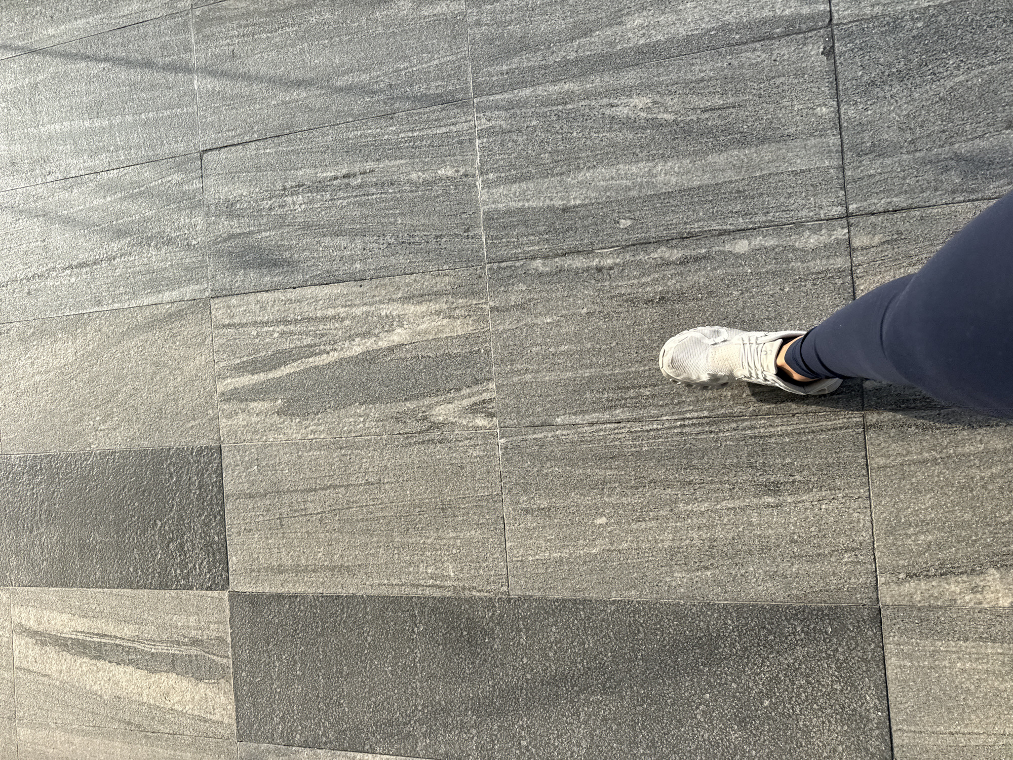 The image captures an overhead view of a person's leg and foot on a tiled surface, likely in Puebla, Mexico. The person is wearing a white athletic shoe or sneaker with dark blue leggings, and their foot is positioned as if in mid-stride or about to take a step.
The ground is covered with rectangular tiles laid in a consistent grid pattern. There are two distinct types of grey tiles used interchangeably. Some tiles exhibit a light to medium grey base with prominent horizontal streaks of white and darker grey, suggesting a natural stone or marble-like pattern with a textured finish. Other tiles are a uniform dark grey with a finely granular, speckled texture. This alternating pattern of streaked and granular tiles creates a visually dynamic ground surface.
The lighting indicates it is daytime, with soft shadows cast across the tiles, suggesting clear and sunny weather, possibly during the mid-morning or mid-afternoon. There are no additional objects, discernible text, or explicit location cues visible within the frame. The scene focuses on the interplay between the pedestrian's movement and the distinctive tiled ground.
