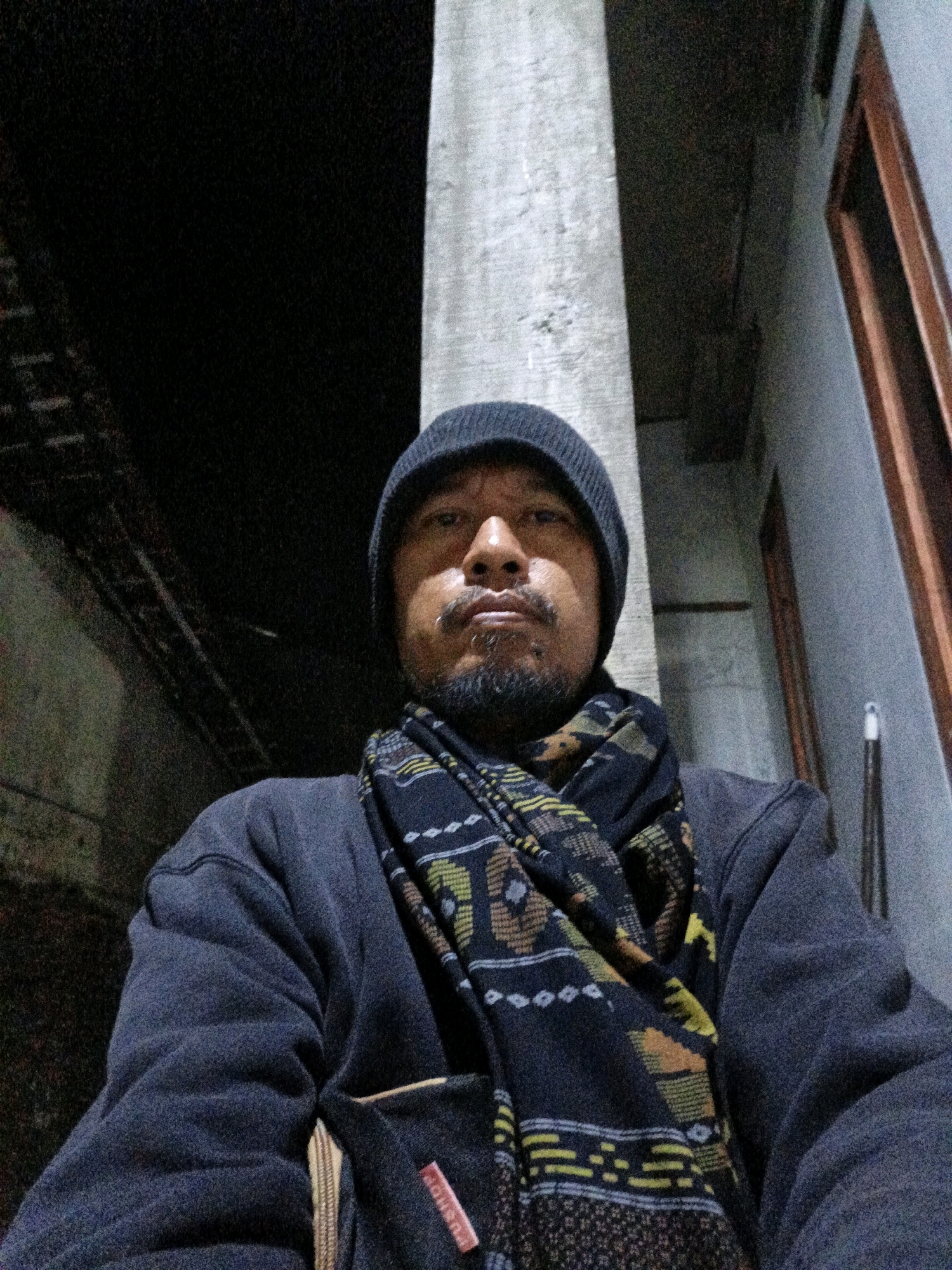 A man wearing a dark beanie, a patterned scarf, and a dark jacket is pictured from the chest up. He has a goatee and a pierced lip. He is outdoors at night, possibly in the city of Tanahbaya, Indonesia, with a dark background and a concrete pillar to his right. The image is dimly lit, with a slight upward angle.