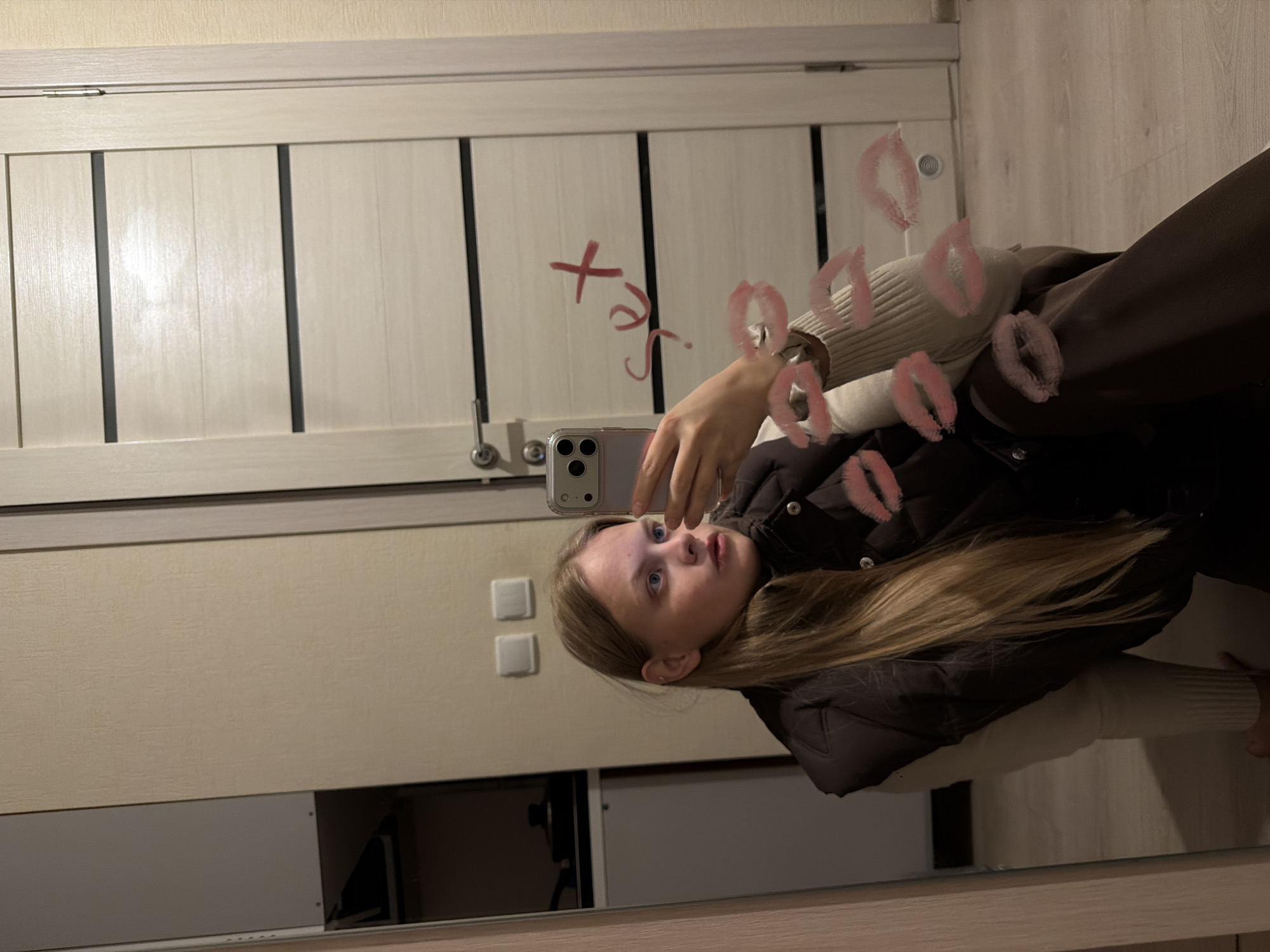 The image is a tilted mirror selfie of a young woman in an indoor setting. The primary subject is a young woman with long, straight blonde or light brown hair, fair skin, and blue eyes. She is wearing a dark puffer jacket over a light-colored, possibly cream or white, ribbed sweater. Her expression is attentive and slightly curious, with her gaze directed upwards and to her left (from the viewer's perspective). She holds a silver smartphone, likely an iPhone, to capture her reflection.
The mirror itself is a notable object, adorned with several bright red lipstick kiss marks. There is also red lipstick writing on the mirror, which appears to spell "xoxo" or similar playful text.
The reflected background reveals an interior space. In the upper portion, a light-colored interior door with three dark vertical decorative strips and a silver doorknob is visible. Above the door, there's a light-beige wall with a subtle texture. Below the door, the wall continues, featuring two square white light switches. The very bottom of the mirror reflects a dark, possibly wooden, surface that could be a desk or dresser, with some obscure dark objects visible within its reflection. The mirror itself is framed by a light wooden border at its base.
The activity taking place is the woman taking a mirror selfie. The scene is lit by artificial indoor lighting. The image is rotated approximately 90 degrees counter-clockwise. The provided location context is Izhevsk, Russia.
