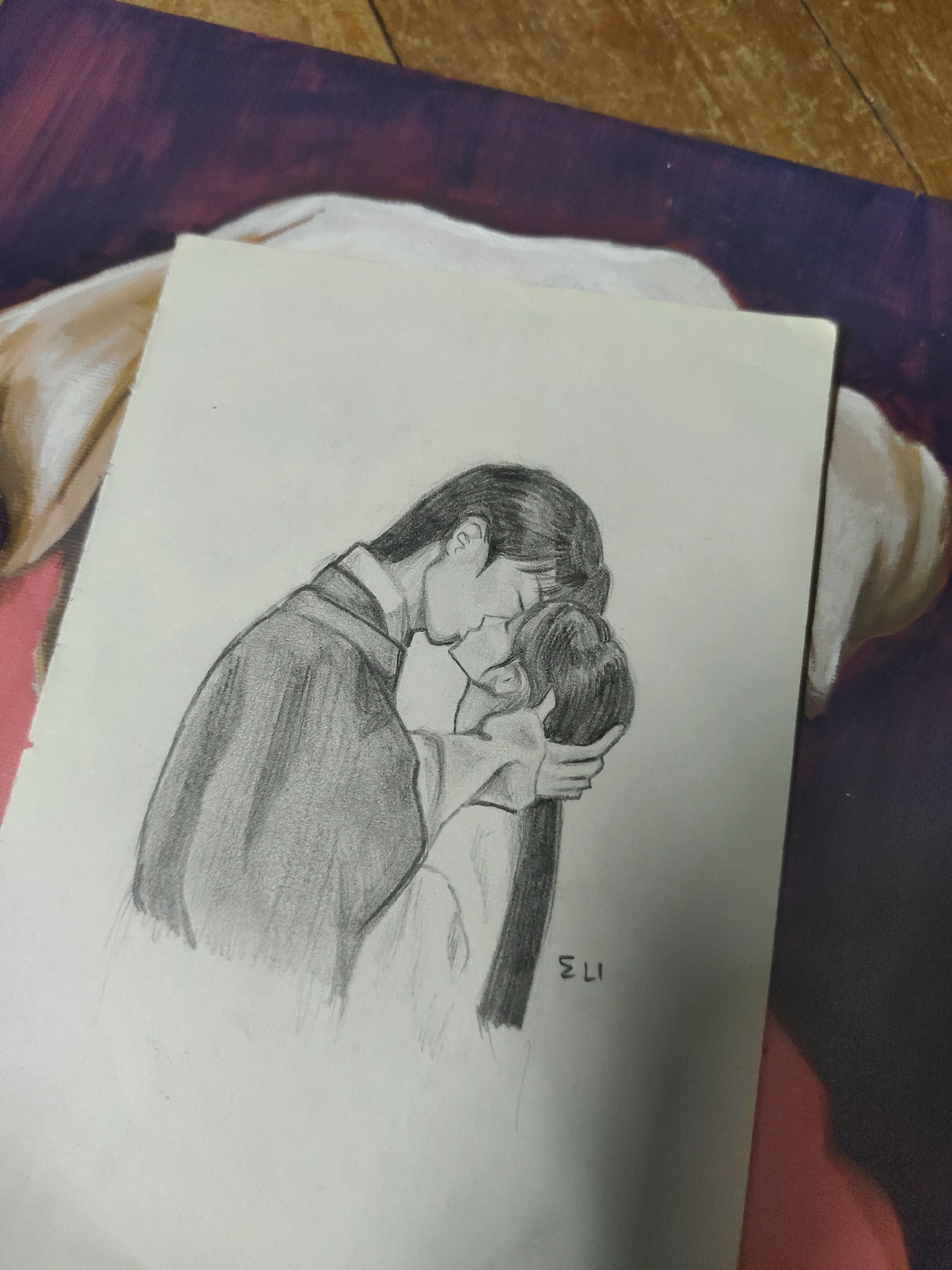 The image displays a graphite pencil sketch on a rectangular sheet of white paper, positioned diagonally across the frame. The sketch depicts two human figures in an intimate embrace, their heads tilted towards each other with lips touching, indicating a kiss. The figure on the left, appearing taller, is rendered in a dark, shaded garment resembling a traditional robe. The figure on the right has long, dark hair and is embraced by the left figure, whose hand gently holds the back of their head. The initials "ELI" are handwritten in the lower right corner of the sketch. The paper rests upon a larger, textured surface, possibly a fabric mat or tapestry, exhibiting mottled patterns in shades of deep purple, brown, and light beige. Portions of a light brown wooden surface are visible in the background, underneath and around the patterned mat. No discernible information within the image indicates a specific location, including Davao City, Philippines.