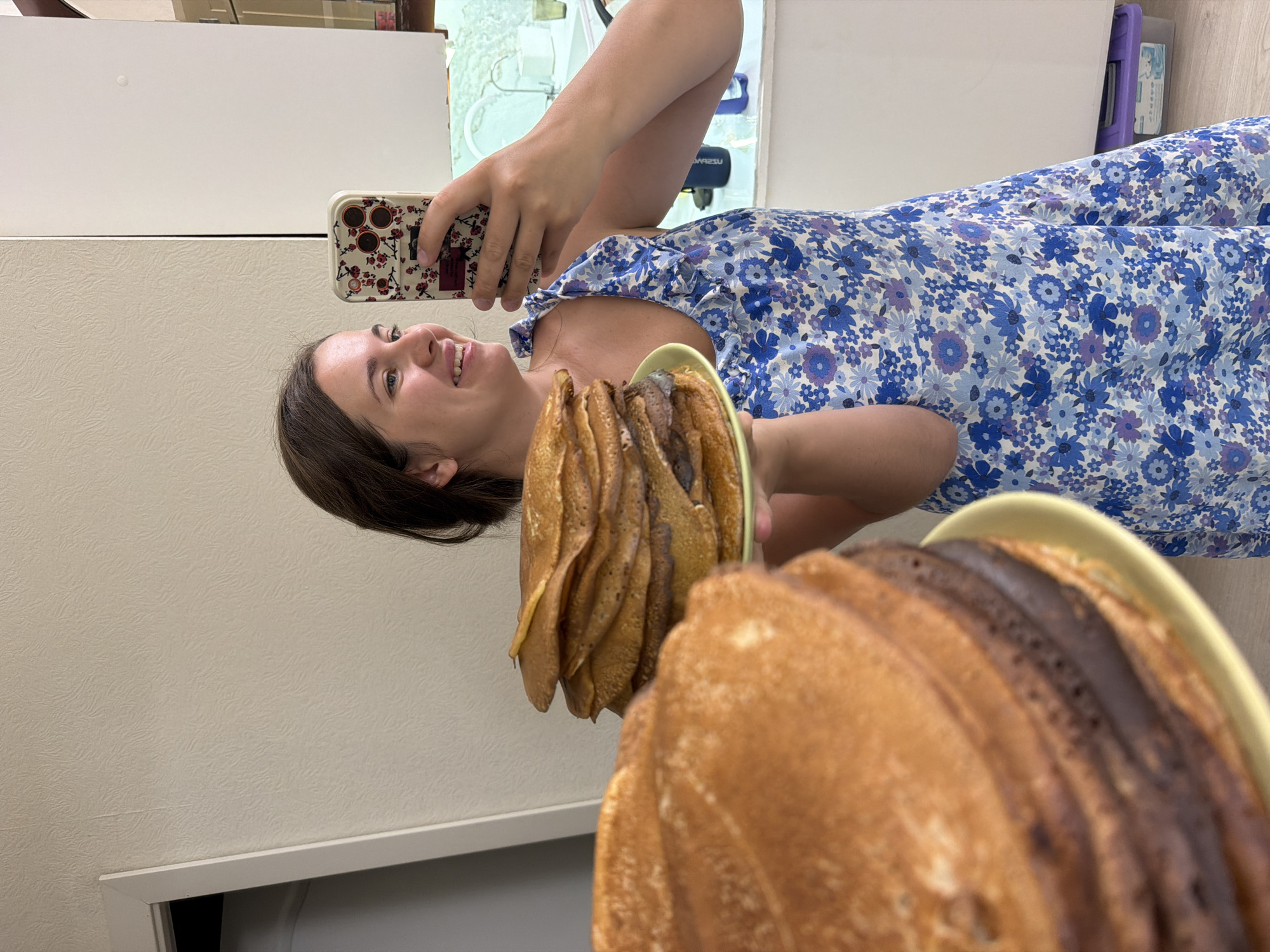 In a kitchen, a woman with brown hair wears a blue floral dress. She is holding a phone in her right hand and taking a selfie. In her left hand, she holds a yellow plate with a tall stack of crepes. Another stack of crepes is in the foreground, slightly out of focus. The background shows a white door frame and a textured wall.