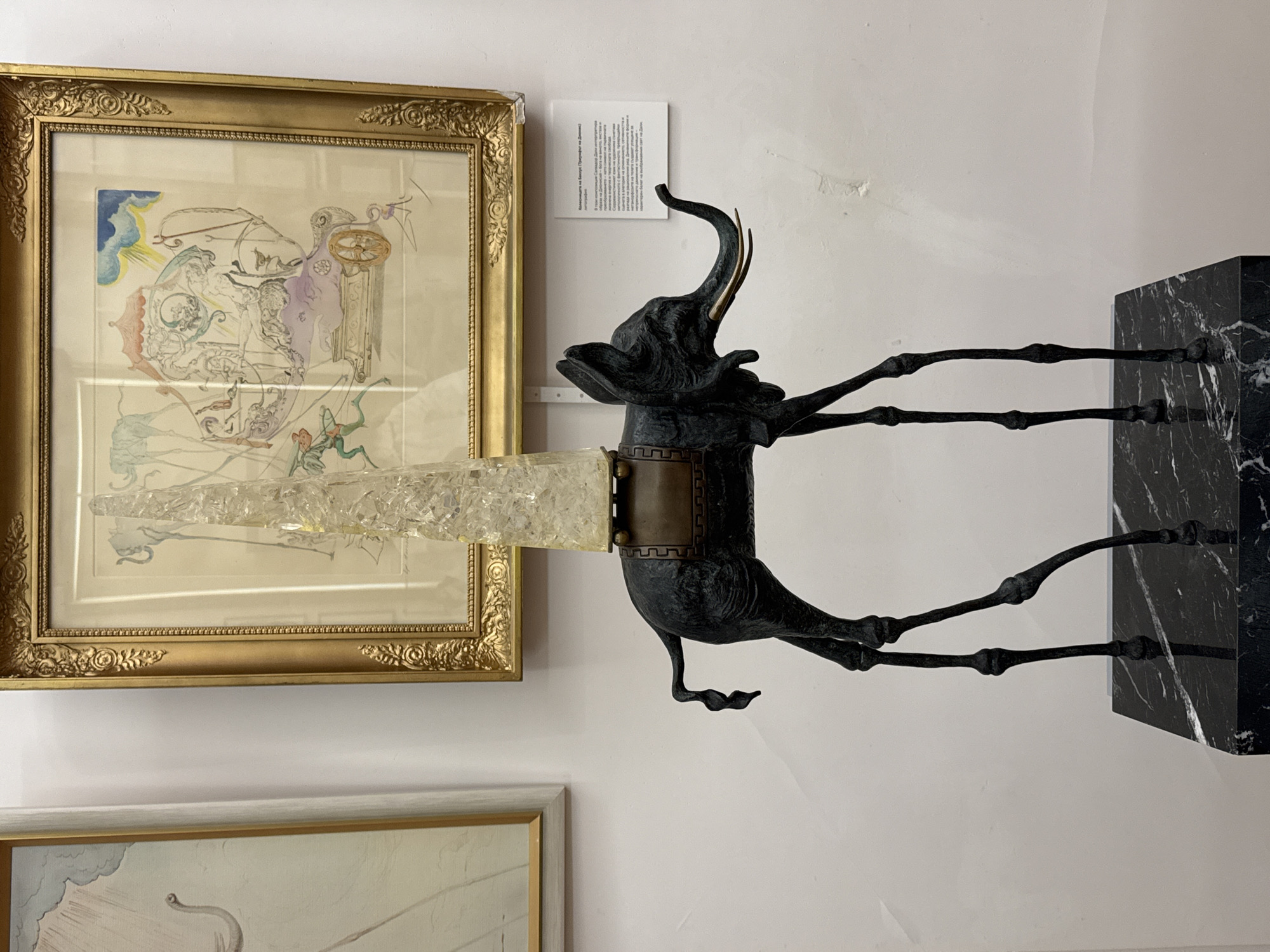 A bronze sculpture of an elephant with impossibly long, thin legs stands on a black marble pedestal. The elephant is dark in color and has a saddle-like structure on its back, topped with a tall, clear, faceted crystal obelisk. Behind the sculpture, a framed artwork hangs on a white wall. The artwork appears to be a surrealist drawing or painting in a gold frame, depicting elephants with long legs, a chariot, and figures. To the left of this artwork, another framed piece is visible, partially cut off by the image border, also appearing to be a drawing with a pale background and light-colored elements. A small white informational plaque is attached to the wall to the right of the elephant sculpture. The overall scene suggests an indoor exhibition space, likely a museum or gallery. The city of Pernik, Bulgaria, is not visually depicted or implied in this image.