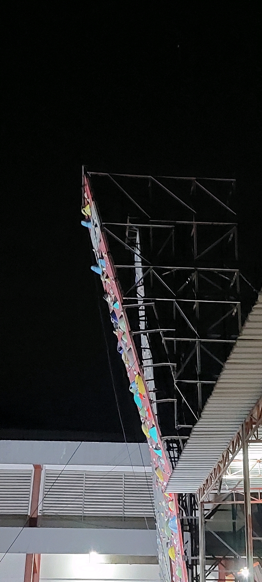 An outdoor, night-time, low-angle, full shot of a climbing wall with colorful handholds attached to a red surface. The wall is angled upwards and to the right, with a large metal structure behind it. To the right of the wall, a silver-colored, sloped roof structure is visible. In the background, a building with louvred windows can be seen. The scene is dark, with the climbing wall and roof being the only illuminated structures. The sky is completely black, with a small, bright spot in the upper center.