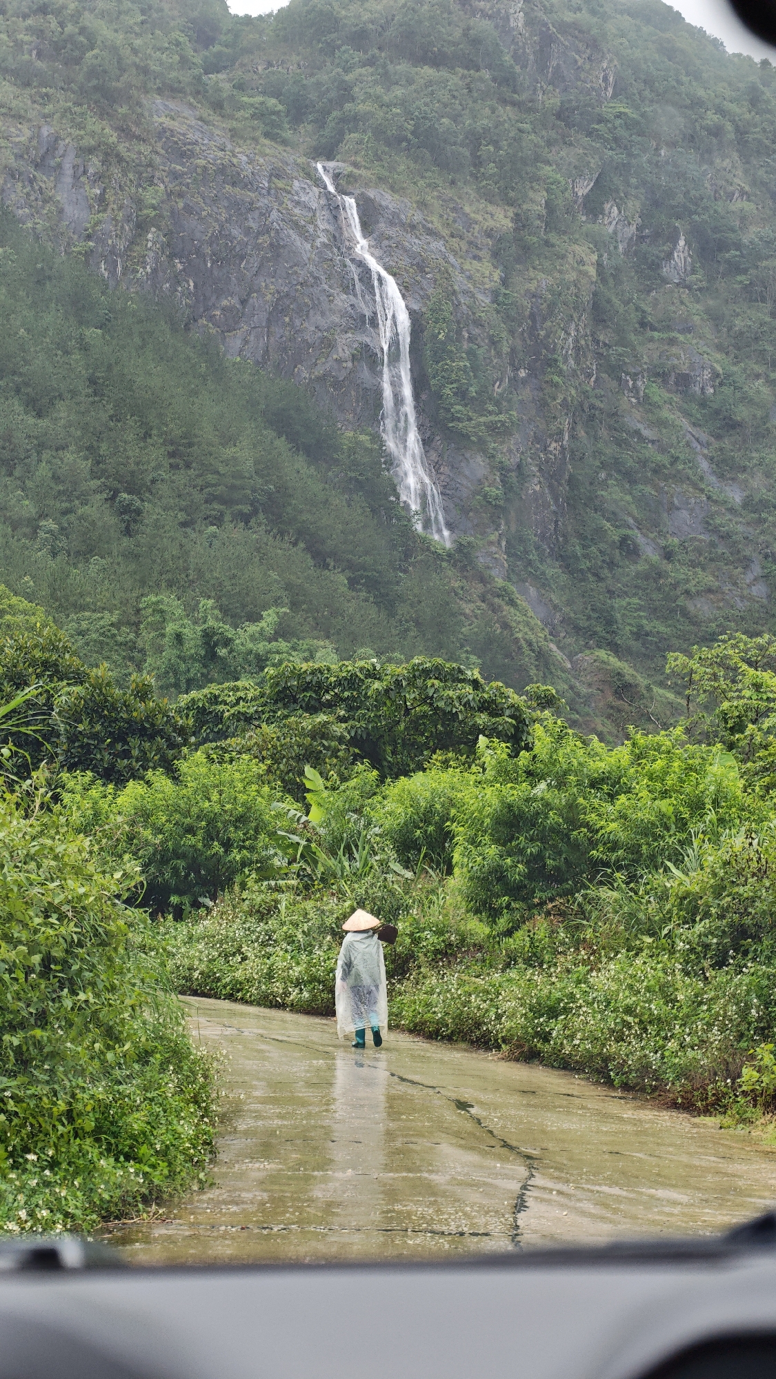 A long shot depicts a person walking away from the viewer down a paved road. The individual wears a traditional conical Asian hat and a translucent poncho, indicating a rainy environment. The road is wet and cracked, with green vegetation lining both sides. In the background rises a mountain with a cascading waterfall. The scene is likely located in Hồ Thầu, Vietnam, given the traditional hat and mountainous environment. The person carries something over their shoulder. The scene appears to be viewed from inside a vehicle.