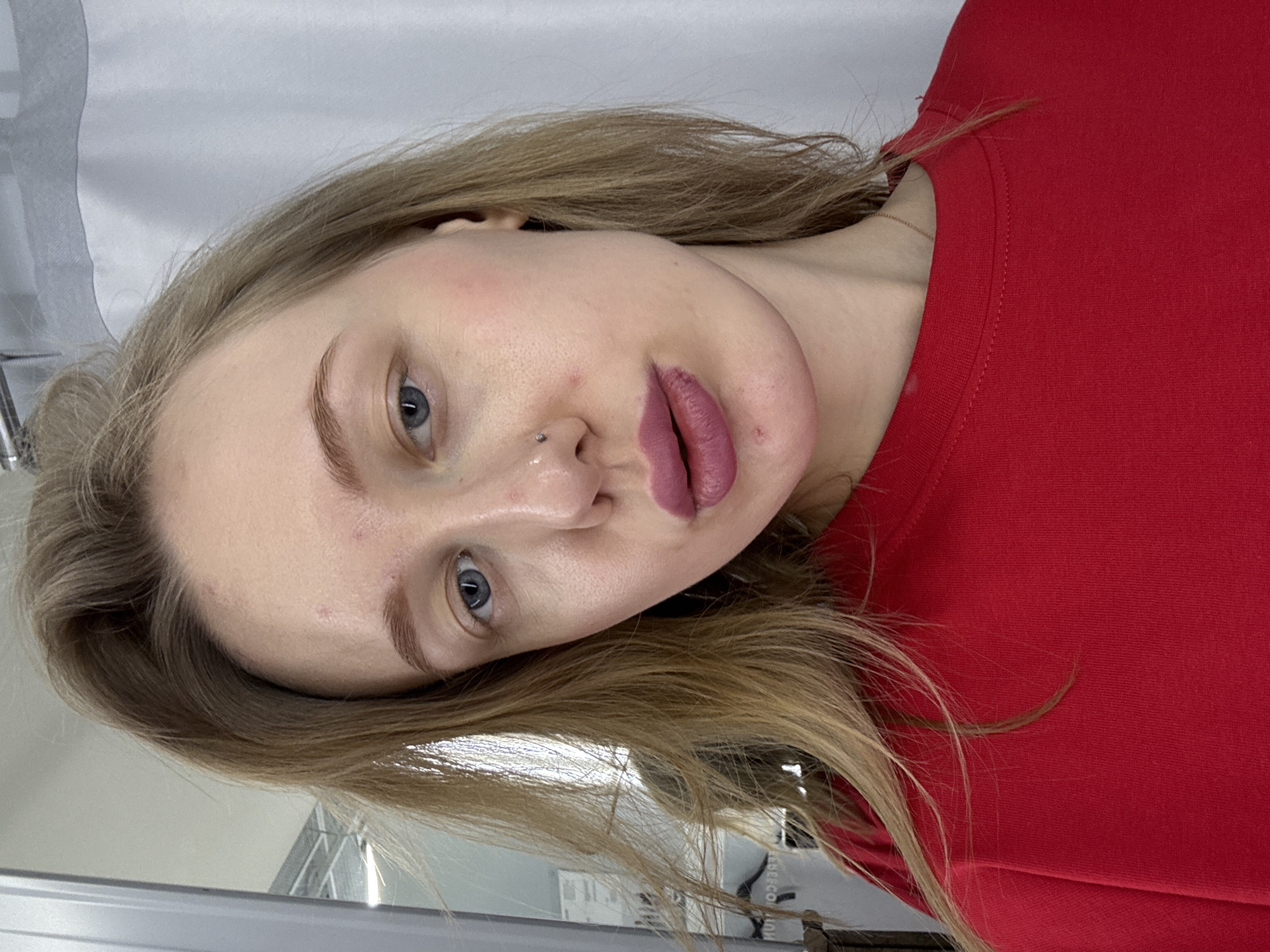 A woman with blonde hair, blue eyes, and fair skin is wearing a red top. She has a nose piercing on her left nostril and is looking directly at the camera with a slightly tilted head. The background appears to be a plain wall or curtain. The image doesn't provide enough context to determine a location in Rostov-on-Don, Russia, or to infer specific actions or interactions.
