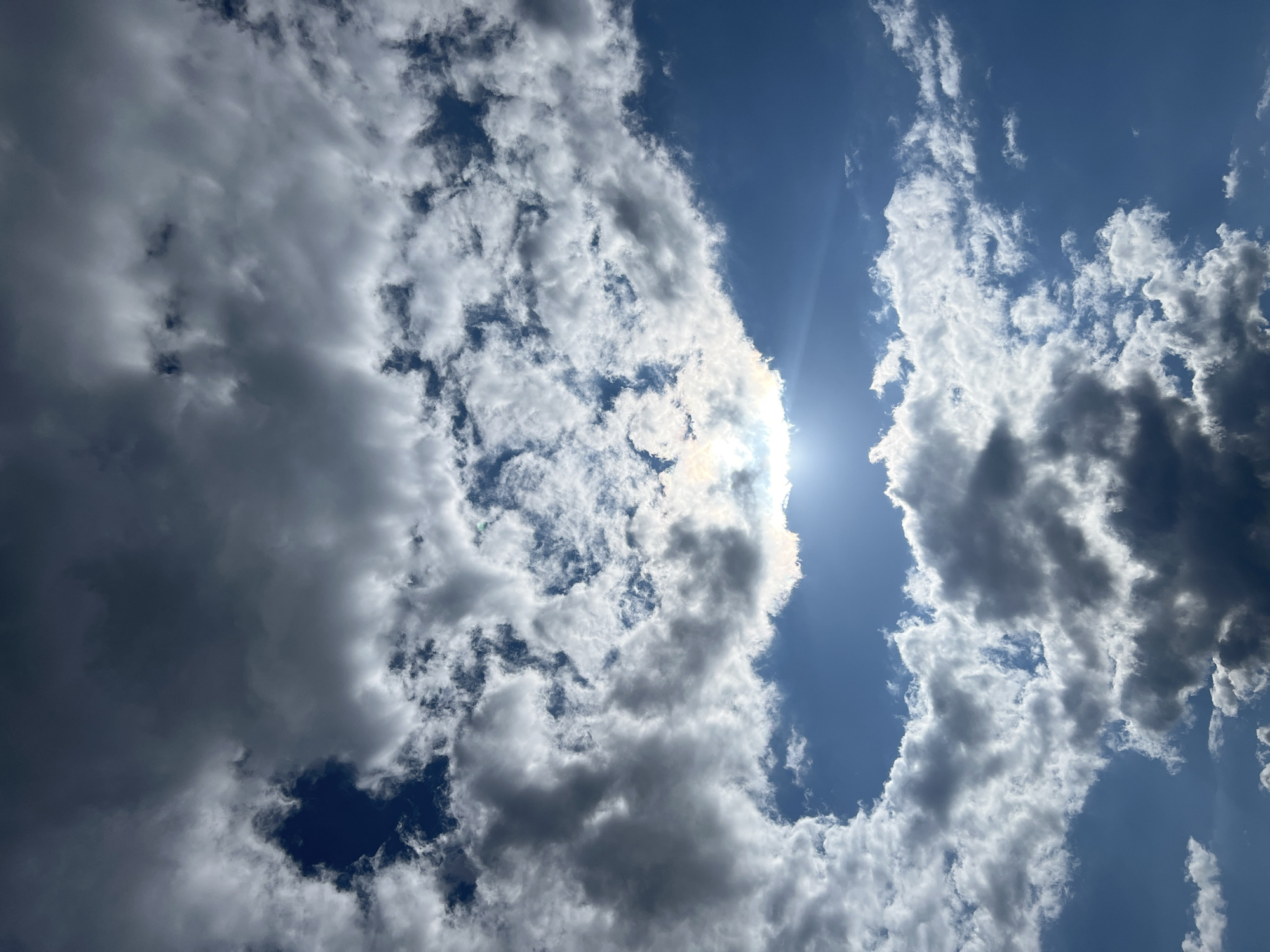 A vertical, low-angle shot captures a dynamic sky over Sobishchytsi, Ukraine, filled with dramatic, varied cloud formations. The bright sun peeks through a large, bright cloud mass in the upper center, casting rays and creating a halo effect. Below this, darker, more ominous clouds drift across a clear, vibrant blue sky. The clouds exhibit a range of textures, from wispy and thin to dense and shadowed, suggesting a mix of weather conditions. The image is devoid of any visible structures, people, or ground-level objects, focusing solely on the atmospheric phenomena.