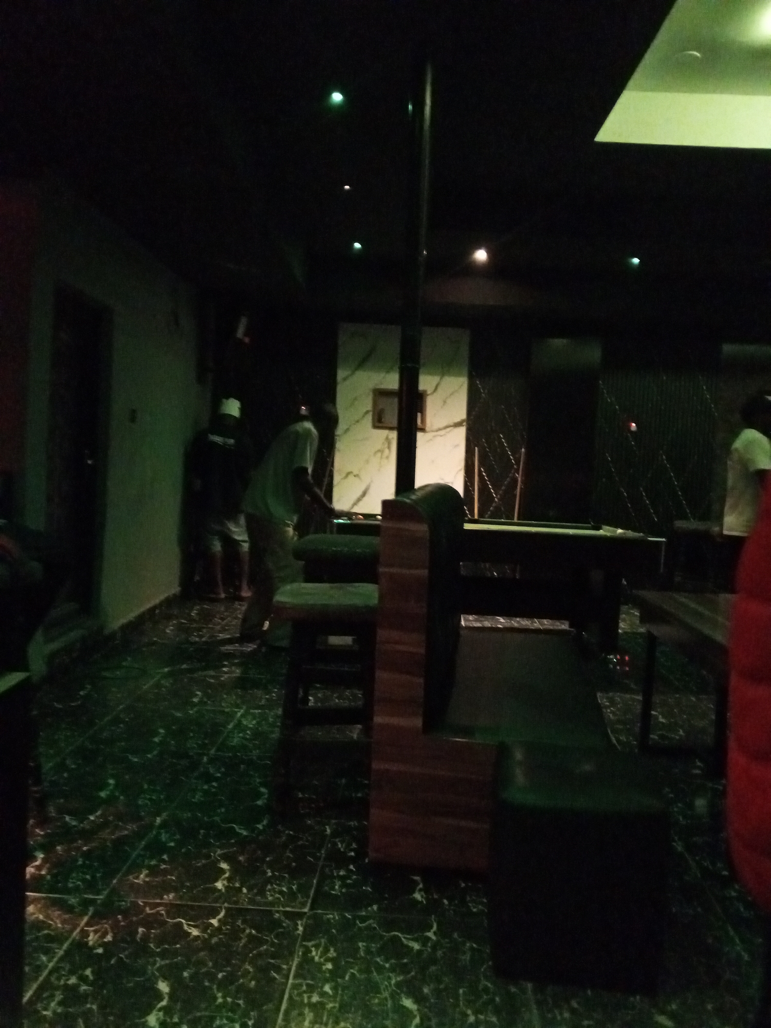 The image depicts the interior of a dimly lit establishment, likely a bar or lounge, with a pool table as the central focus. The floor is tiled with a dark green and white marbled pattern, reflecting the ambient green lighting. Several individuals are present. On the left, two men are standing near a wall, one wearing a white cap and the other a white shirt. To the right of the pool table, another individual in a light-colored shirt is seen bending over. The pool table itself is dark and appears to be in use, with cues leaning against a rack in the background. The overall atmosphere is dark and somewhat subdued, with spotlights or ceiling lights casting pools of light. The walls are a mix of dark surfaces and textured panels, with some framed artwork visible.
