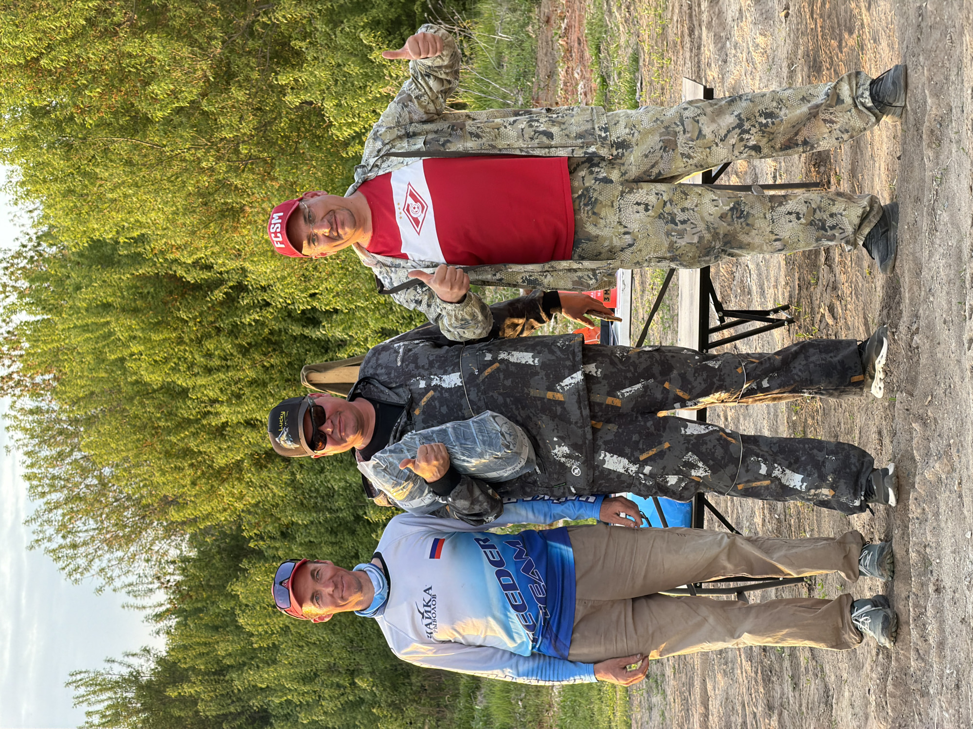 Three men are standing outdoors.
The first man, closest to the viewer, is wearing a long-sleeved, blue and white shirt, khaki pants, and a red baseball cap.
The second man is wearing a black jacket with grey accents and black pants. He is also wearing sunglasses and a black baseball cap. He is holding a bundled object wrapped in clear plastic.
The third man is wearing a camouflage jacket, a red t-shirt with a white logo, and a red baseball cap. He is giving a thumbs-up.
All three men are standing near a folding table.
The background is a combination of trees and a dirt ground.
Based on the "FCSM" on one of the caps, it is likely the location is in the city of Krasnyi Bor, Russia.