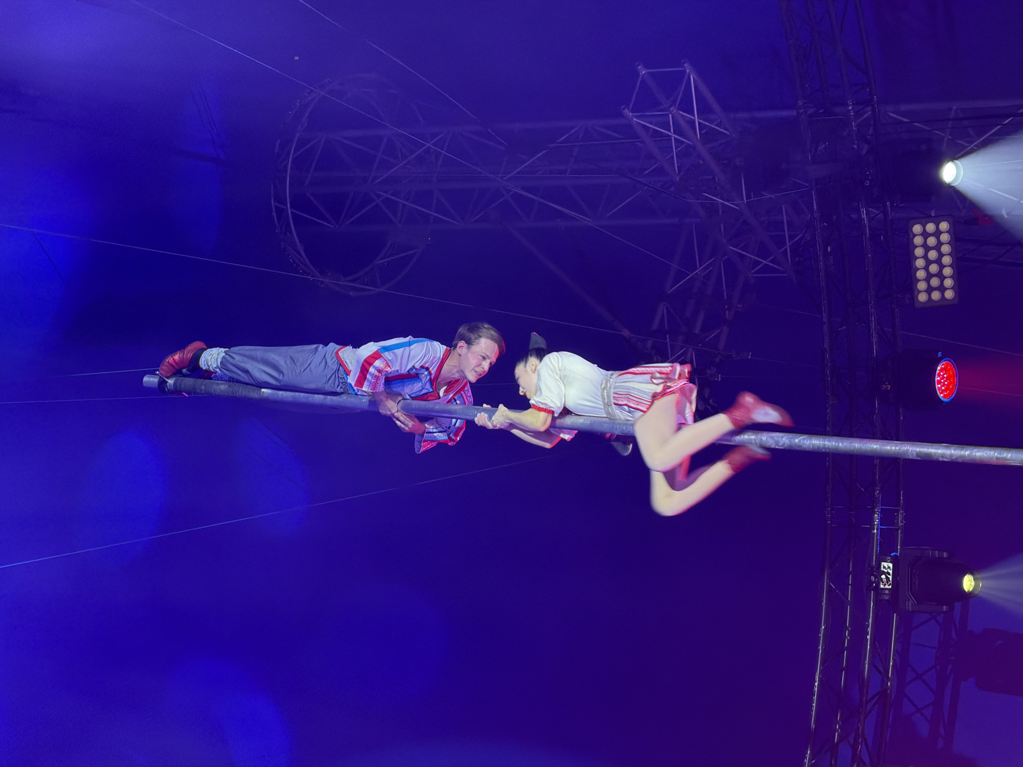 Two acrobats, a male and a female, are suspended mid-air on a horizontal bar, performing an act. The male acrobat, on the left, wears a red, white, and blue striped costume with red shoes, and he is lying on his stomach with his hands gripping the bar. The female acrobat, on the right, is positioned above and facing him, also lying on her stomach. She is dressed in a white and red striped outfit with a red bow in her hair and red shoes. Her hands are reaching out towards his, appearing to be holding onto his wrists or arms. Their bodies are positioned in a way that suggests a moment of balance and connection in their performance.
The setting appears to be a circus tent or a performance venue, indicated by the blue backdrop, rigging structure above, and spotlights illuminating the stage. One spotlight shines brightly from the top right, casting a beam of light. Another lighting fixture with multiple bulbs is visible to the right of the center. The overall lighting is dramatic, with deep blues and bright highlights. There are no visible indications of time of day or weather. The focus of the image is on the acrobats and their precarious act, highlighting their skill and the tension of the performance. There is no visible text in the image.