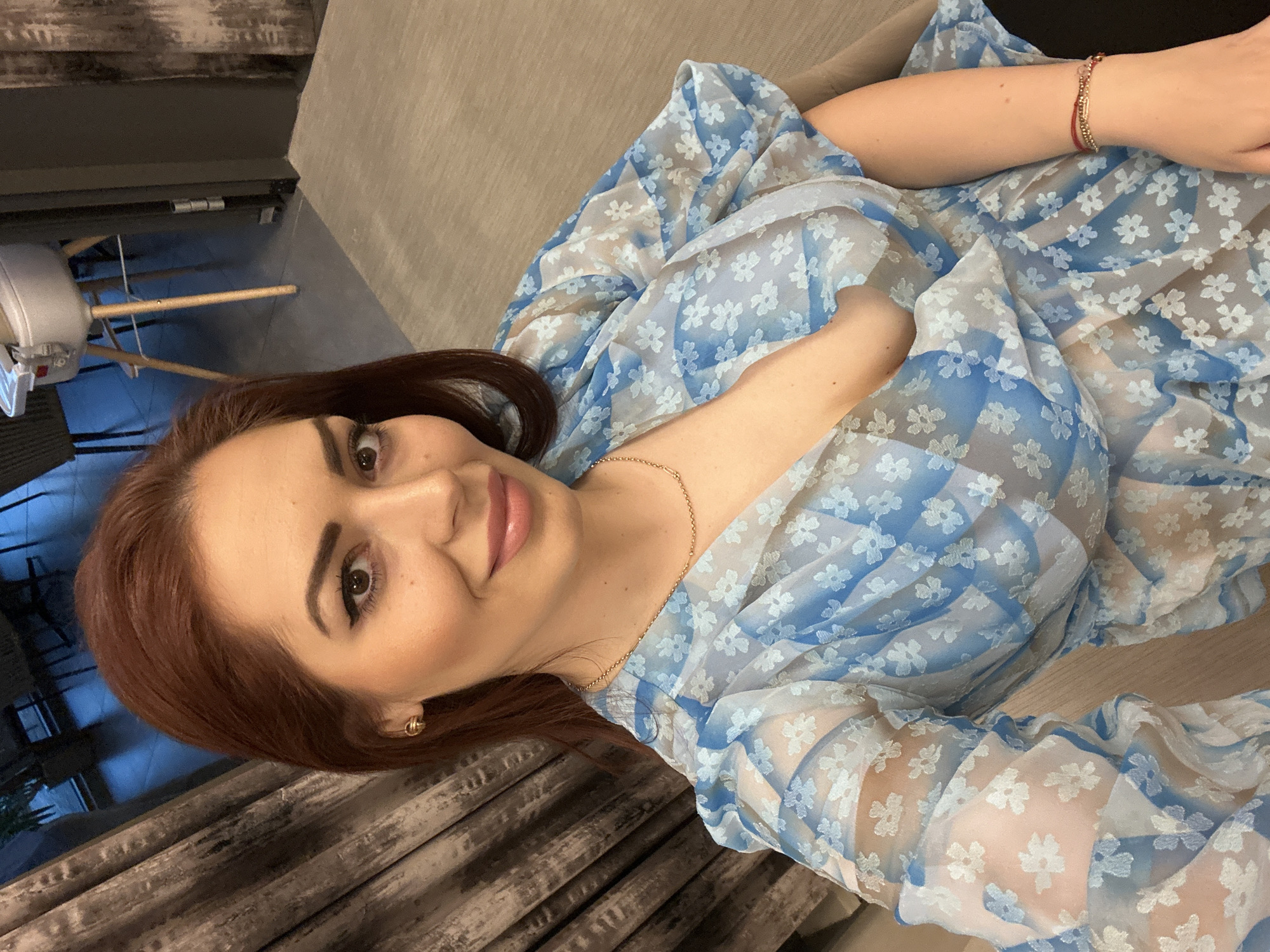 A woman with auburn hair, fair skin, and dark eyebrows and eyelashes is lying on a neutral-toned sofa, possibly indoors. She is wearing a blue and white floral patterned dress and gold jewelry, including a necklace and a bracelet. Her face is slightly turned towards the camera and she is smiling. To the left of the frame, there is a glimpse of a window with visible reflections and a lamp with a tripod stand. The background appears to be part of a residential interior. It is not possible to confirm the location as Kazanlak, Bulgaria, from the image alone.