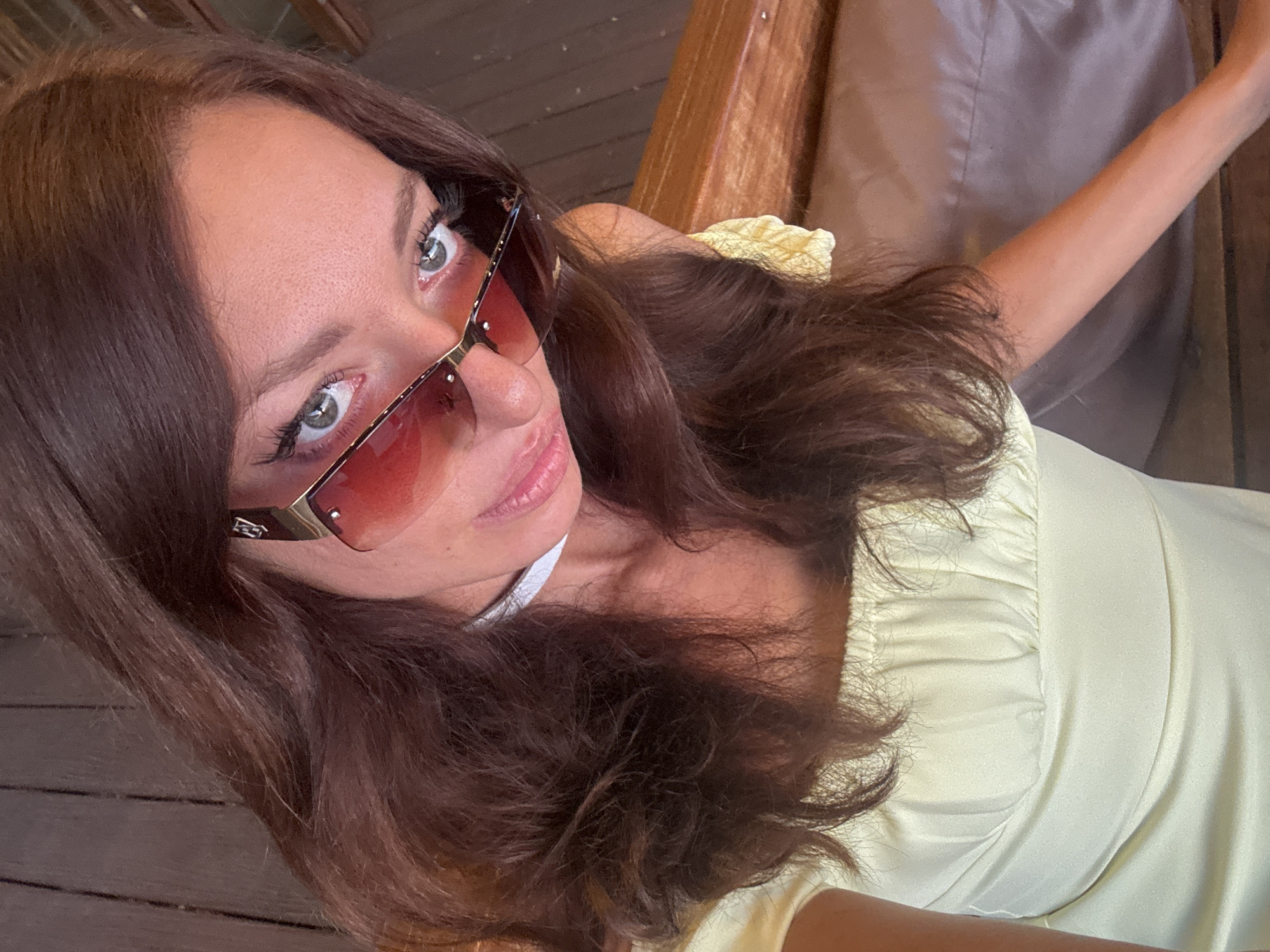 A medium shot shows a woman posing in Baan Plai Laem, Thailand. She is wearing sunglasses with red lenses and a light yellow top with ruffled sleeves. She has brown hair and light eyes. She is looking into the camera with a slight smile. The background includes wooden features of the structure she is in and a light-colored cushion. She is sitting at a table with wooden planks, her right arm extending out.