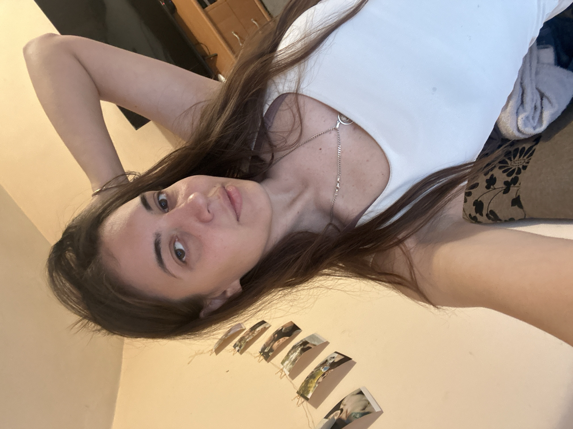 A young woman with long brown hair is posing for a selfie. She is wearing a white tank top and a silver necklace. Her left arm is raised behind her head. In the background, there is a wall with several small framed pictures attached to it, a television, and a wooden cabinet. She is sitting on a patterned couch covered with a gray blanket. The location is unknown and there is no indication of Sirakovo, Bulgaria.