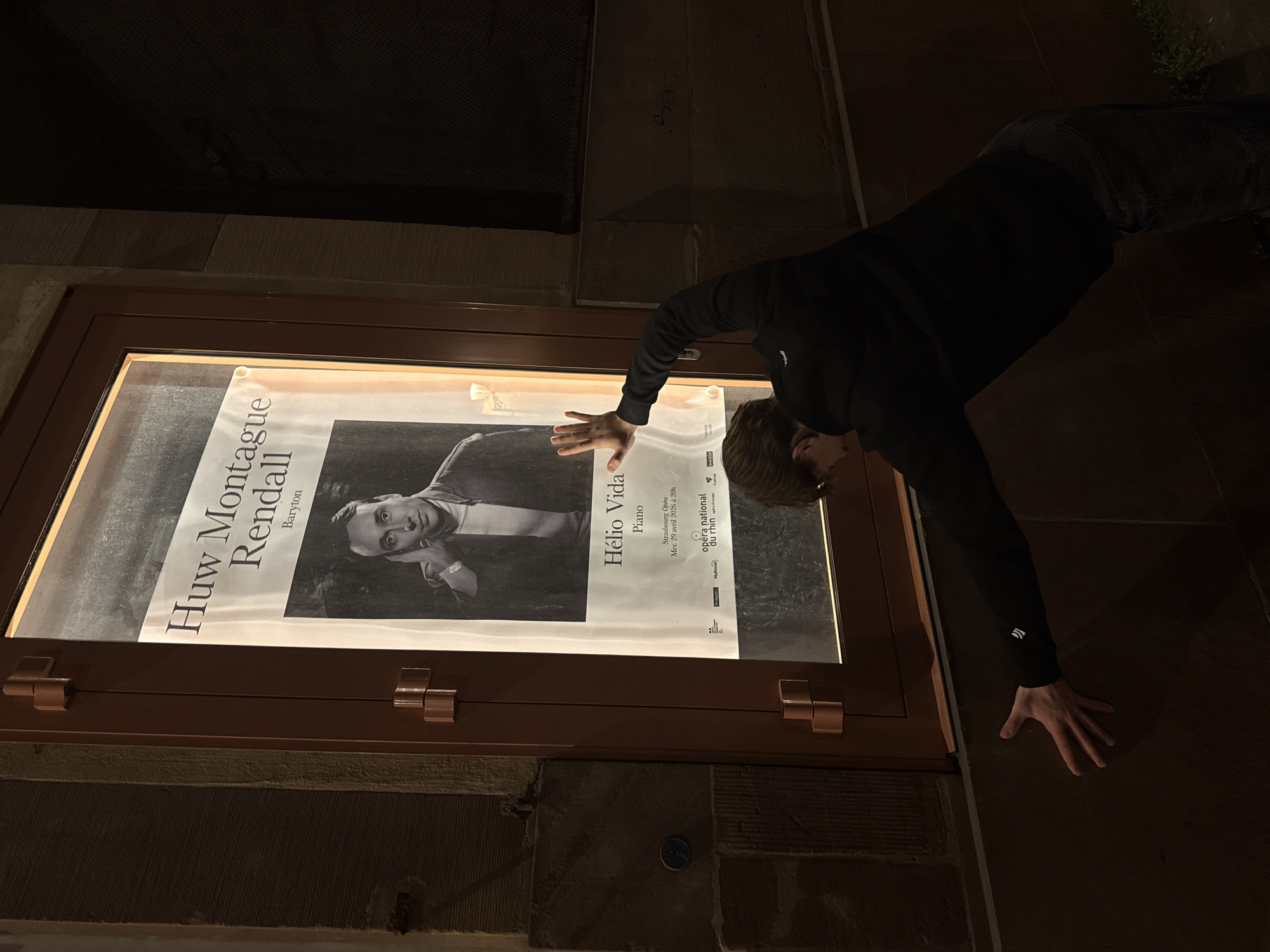 The image shows a person in a black hoodie and dark pants leaning over a poster displayed in a glass-fronted display case. The poster advertises a performance featuring Huw Montague Rendall (Baryton) and Hélio Vida (Piano) at the Strasbourg Opéra National du Rhin on Wednesday, April 29, 2026, at 20:00. The location of the Opera is Strasbourg, France. The person is touching the glass over the poster with their left hand and using their right hand for support on the ground. The background is dark.