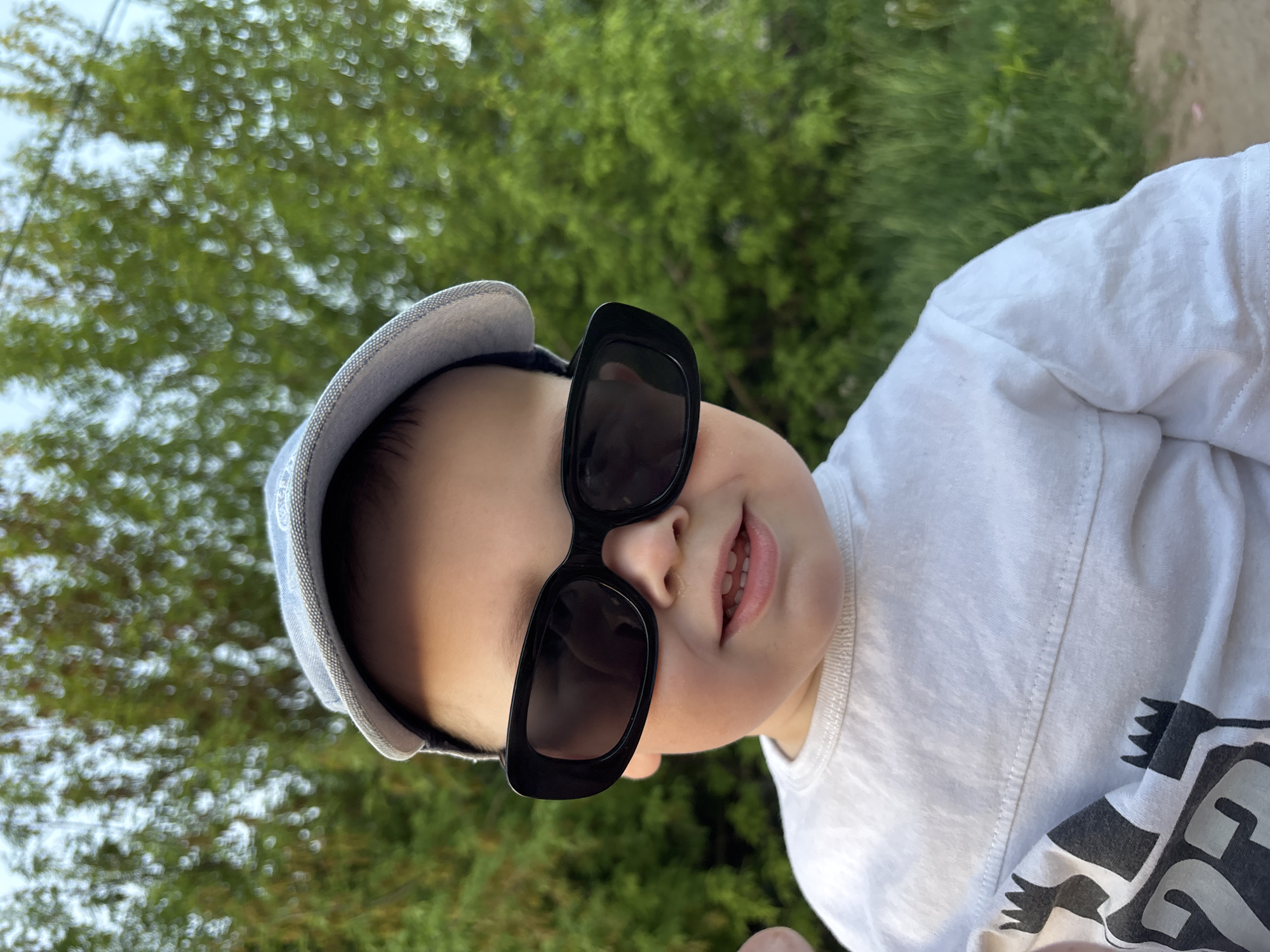 A close-up, tilted shot captures a young child wearing a light-blue cap and oversized black sunglasses. The child has fair skin and a slight smile, revealing a few teeth. They are wearing a white T-shirt with a black and white graphic design. The background is a blurred green foliage, suggesting a natural setting. The photograph is taken outdoors, likely in daylight. The photograph does not indicate the location is in the city of Фоки, Russia.