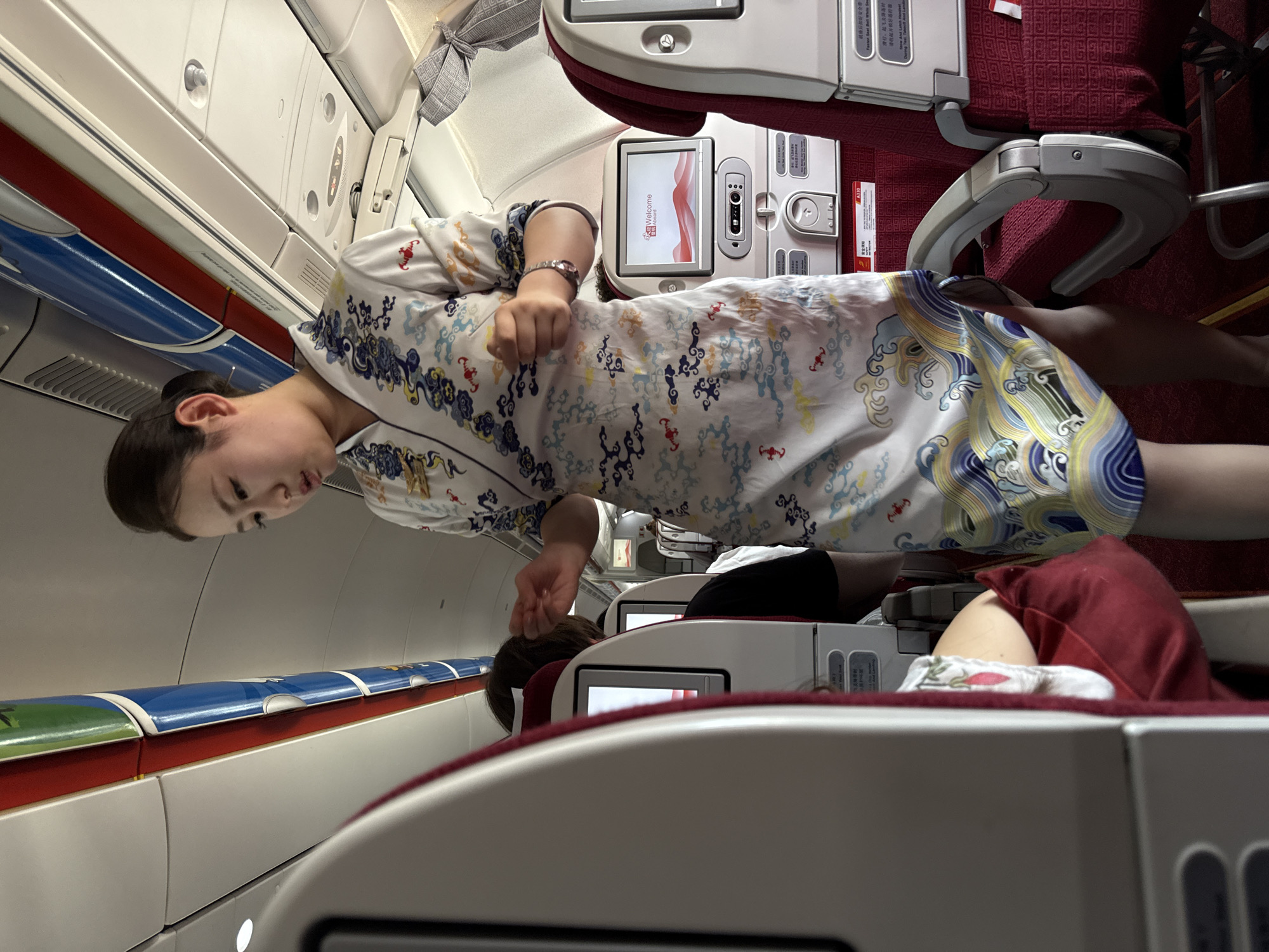 An Asian flight attendant with her hair in a bun is standing in the aisle of an airplane. She is wearing a white uniform dress with a blue and gold cloud and wave pattern. Her right hand is slightly raised, and she appears to be speaking. In the background, there are rows of red airplane seats with attached entertainment screens. One screen displays a "Welcome Aboard" message. The interior of the airplane is visible, including the overhead compartments and lighting. The scene is indoors and appears to be within an airplane cabin, likely in Xiaoshan District, China, given the context of its possible origin.