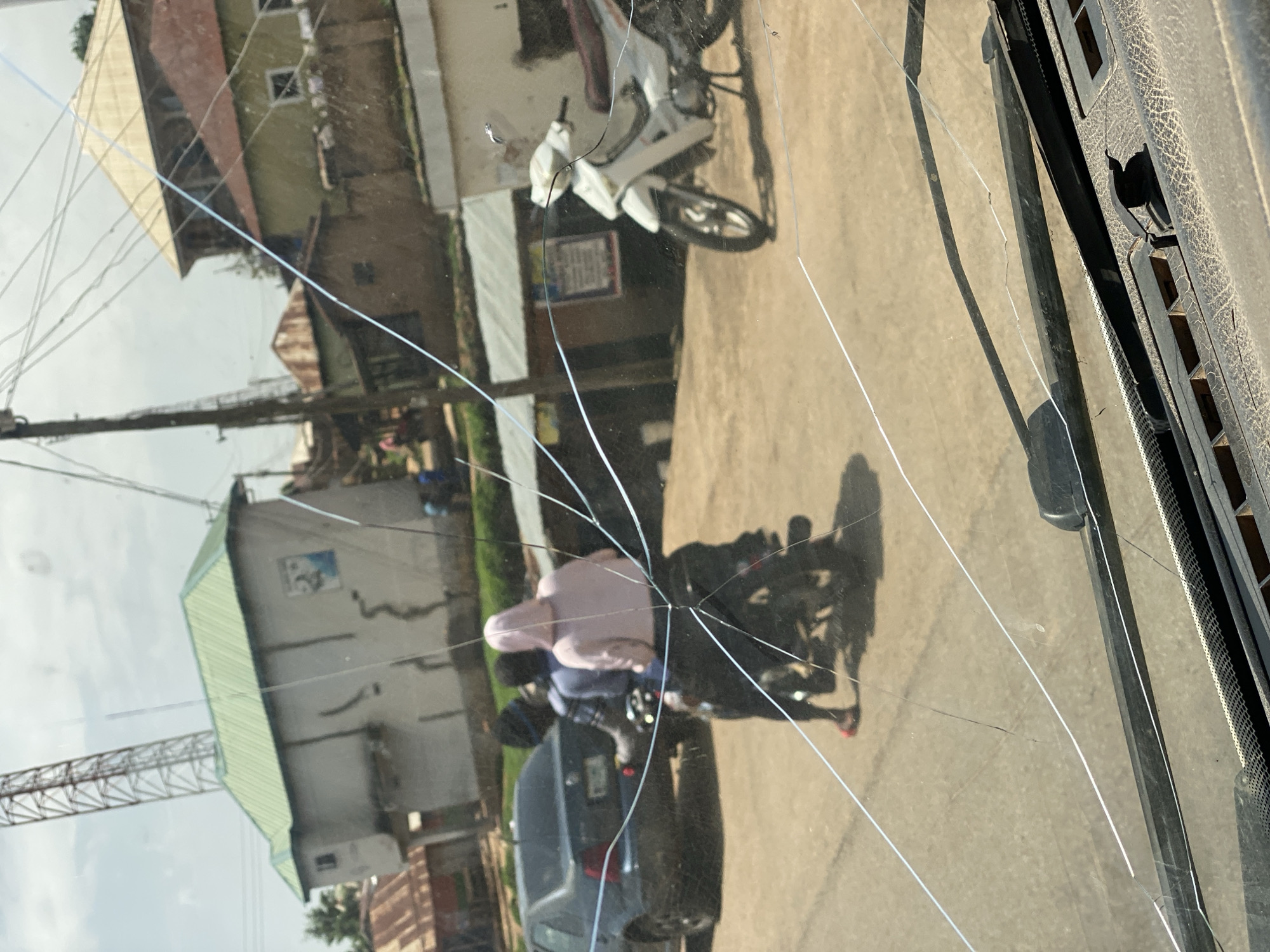 The image captures a street scene in Jos, Nigeria, viewed through a cracked windshield. In the foreground, the dashboard and windshield wipers of a vehicle are visible, with prominent cracks traversing the glass obscuring the view.
The midground shows two individuals riding a motorcycle. The person in the back is wearing a pink hooded garment, while the person in front wears a blue shirt. They are traveling on a paved road. To the left, a dark-colored sedan is parked. Further back, a white motorcycle is parked on the side of the road.
The background reveals a cluster of buildings with various roof types, including a light green corrugated metal roof on a prominent structure and reddish-brown roofs on other buildings. A tall communication tower is visible in the distance. The overall environment suggests an urban or semi-urban setting. The sky is overcast.