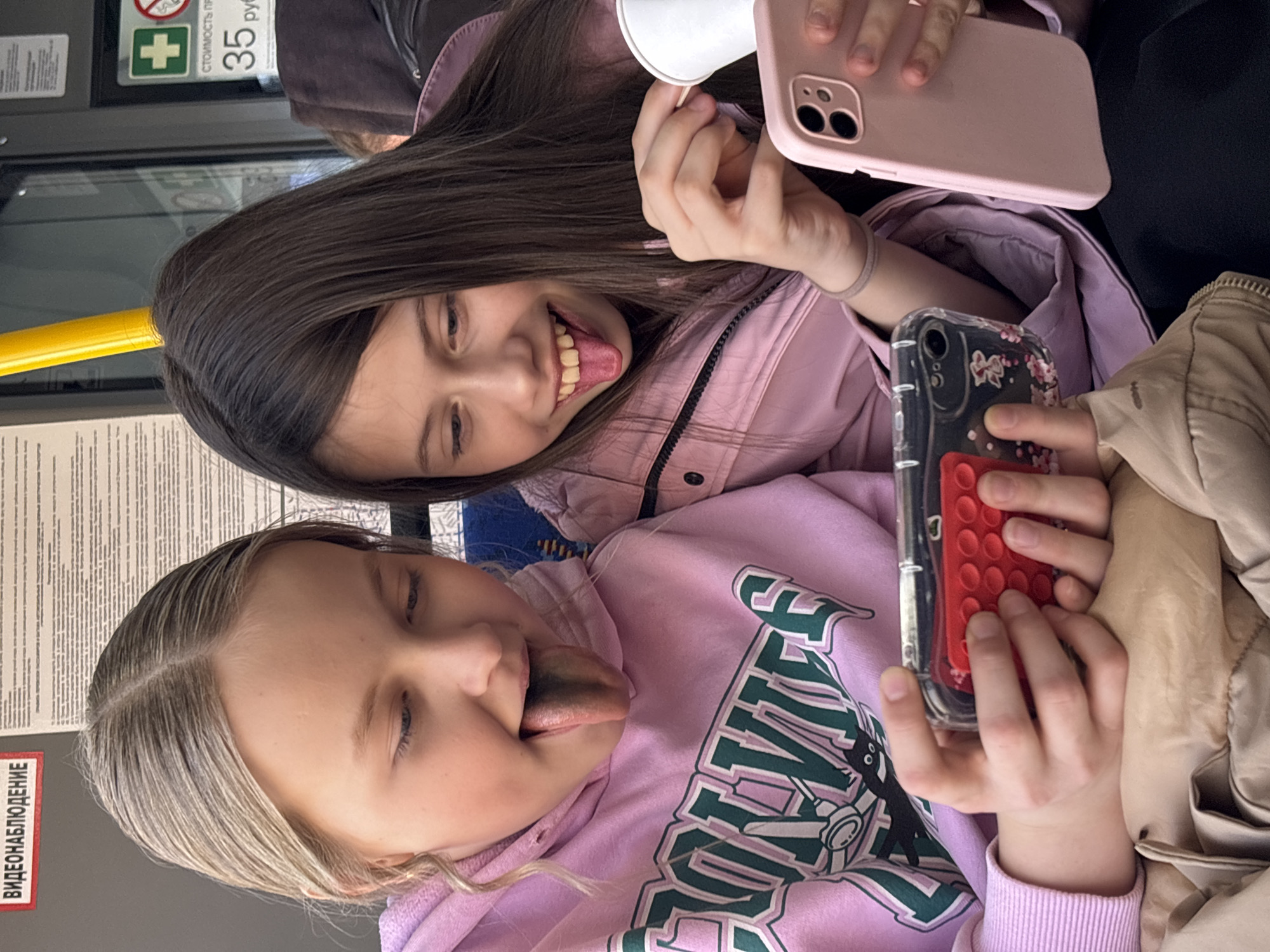 Two Caucasian girls in a public transport vehicle, likely a bus or tram, in Izhevsk, Russia. The younger girl has blonde hair and is wearing a pink sweatshirt with a dark green and white design, and is sticking her tongue out, which is blue. She is holding a cell phone with a transparent case and a red pop socket. The older girl has brown hair and is wearing a light pink jacket. She is also sticking her tongue out and holding a white paper cup and a pink cell phone. They are both looking at the camera, apparently taking a selfie. There are signs in Russian on the vehicle wall, indicating safety information and fare costs. The overall lighting suggests it is daytime.