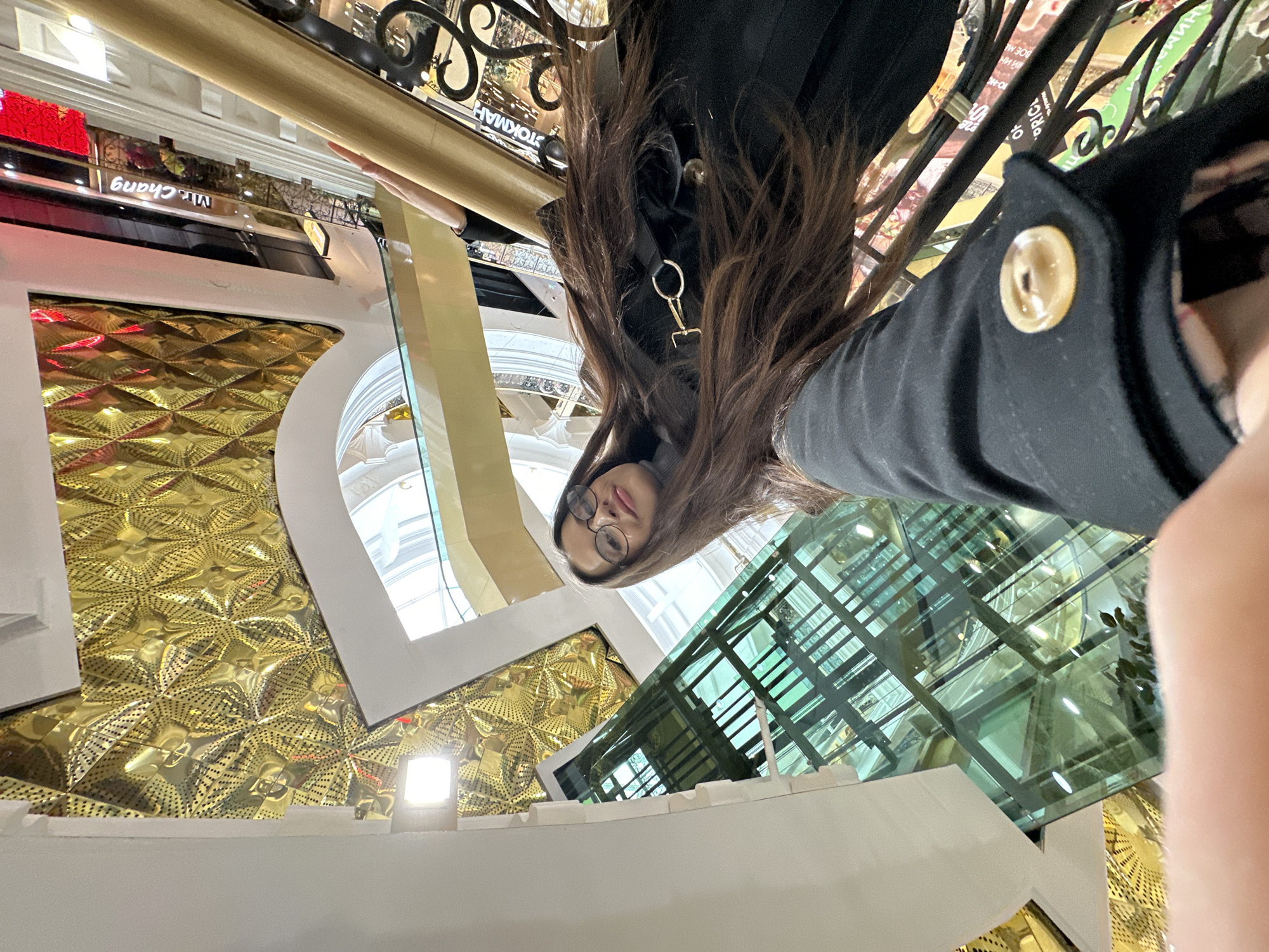 A selfie taken from an unusual angle depicts a woman with long brown hair, wearing glasses and a black jacket with a gold-colored clasp and button. She is positioned in front of a decorative building interior. The background reveals a gold geometric pattern on the walls, large arched windows, and a green glass structure. A sign reading "Mr. Chang" is visible. The setting appears to be a commercial or public space in Yekaterinburg, Russia.