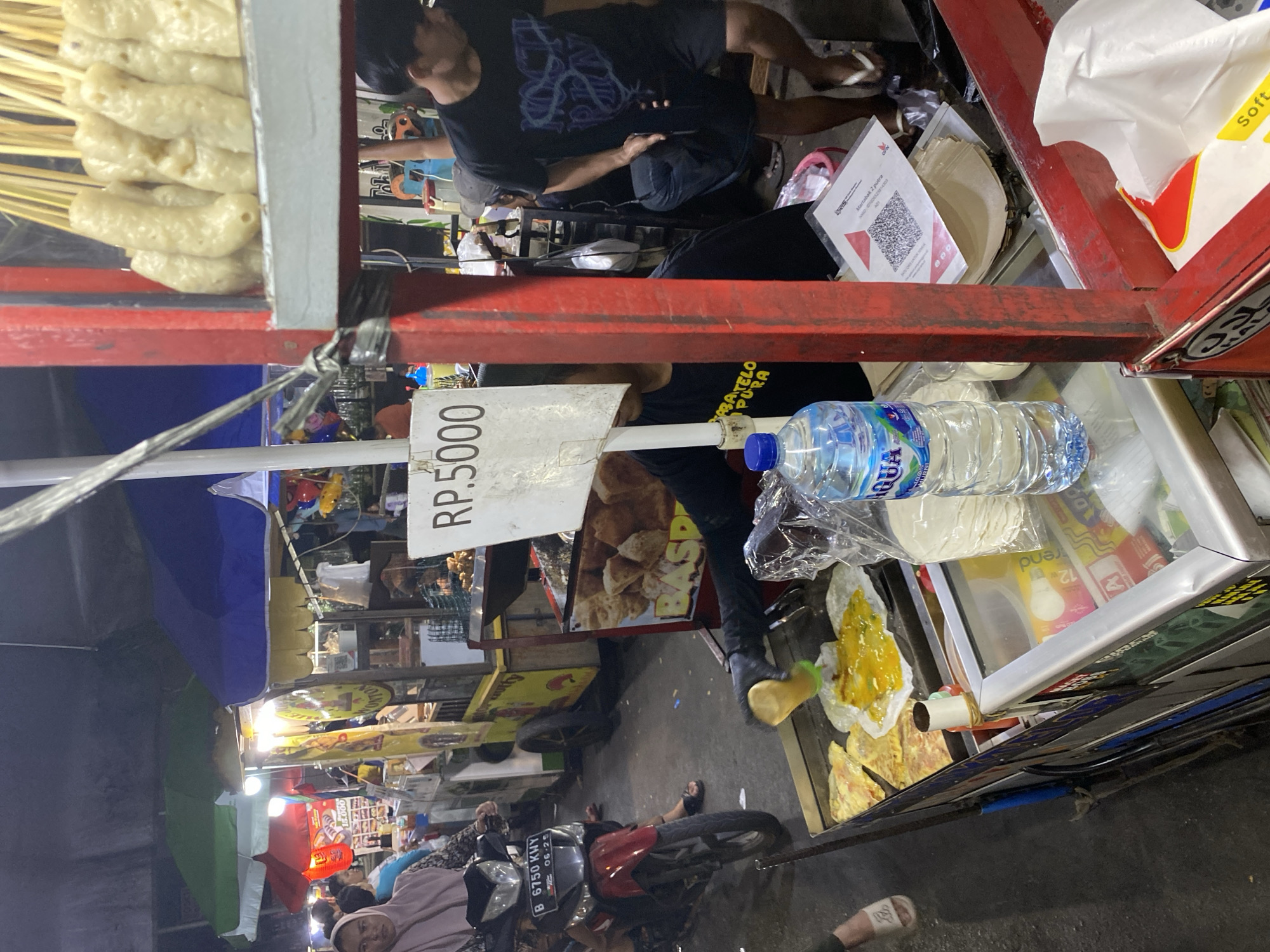 This image shows a street food vendor preparing food at a night market in South Tangerang, Indonesia. A person wearing black gloves is cooking on a griddle, adding a yellow sauce from a bottle onto a thin pancake. Various food items are visible on the griddle and in the display case. In the foreground, a bottle of Aqua water and a stack of thin wrappers are present. Behind the vendor, a sign displays "RP. 5000". Several other people are seen in the background, with a motorcycle parked to the left. The scene is lit by artificial lights, indicating it is nighttime. Umbrellas and food stalls suggest a busy market atmosphere. The overall environment is typical of a vibrant urban street food scene in Indonesia.