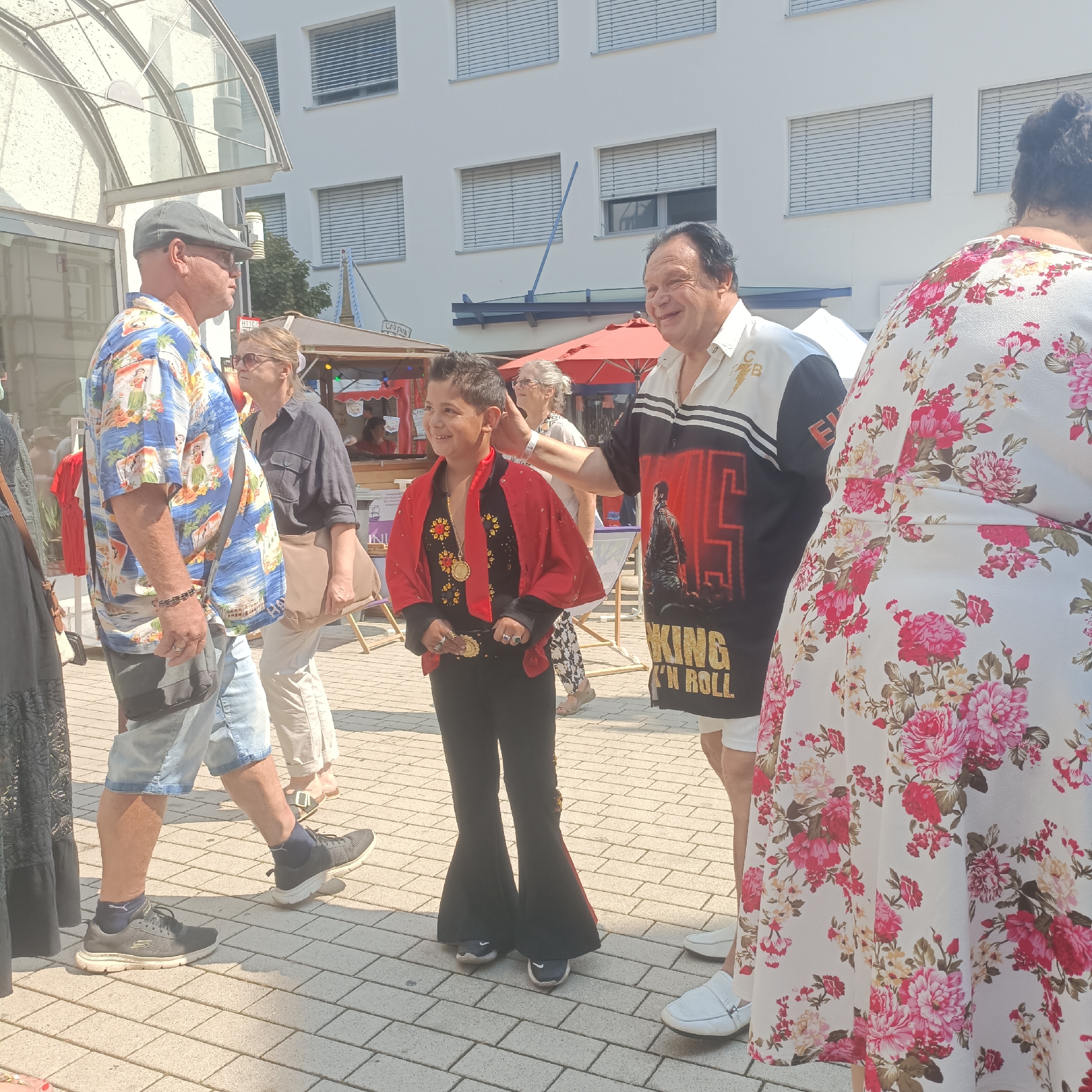 In the city of Bad Nauheim, Germany, a boy dressed as Elvis Presley, complete with a red sequined jacket, gold necklace, and flared pants, smiles as a man in a black and white shirt with "King in Roll" printed on it places his hand on the boy's shoulder. They are standing in a paved outdoor area, possibly a marketplace or street fair, with a white building with blue-slatted windows in the background. Other people are visible, including a man in a Hawaiian shirt and shorts walking by, and a woman in a floral dress. Red awnings and market stalls are also present. The lighting suggests it is daytime and sunny.