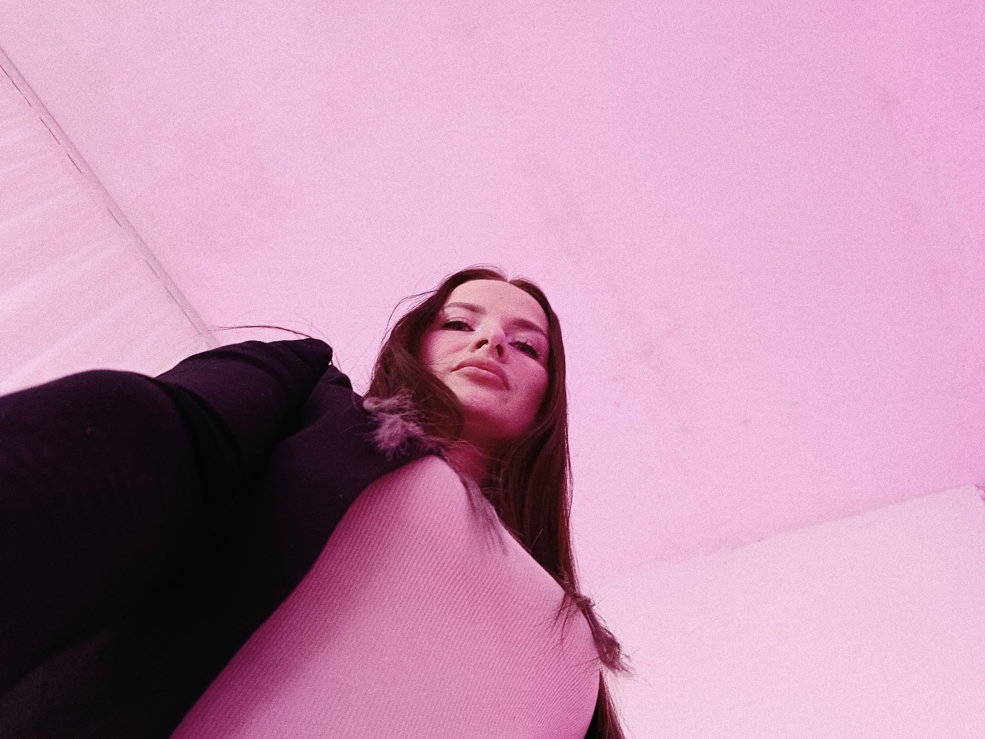 A low-angle indoor shot features a young woman with long, dark hair, looking directly into the camera. Her face and upper torso are visible, positioned in the lower-middle of the frame, with the majority of the image dominated by a plain, pink-hued background that appears to be a ceiling or wall.
The woman is wearing a dark top, possibly a jacket or coat, layered over a lighter, ribbed garment in a pinkish-beige tone. Her expression is neutral to slightly serious, with a direct gaze towards the viewer. The perspective is a worm's eye view, making her appear to loom slightly over the camera.
The overall color palette is dominated by pinks, both from the background and the lighting or filter applied to the image, which casts a warm, monochromatic glow. The background shows a subtle texture, possibly from the surface itself or image noise. No specific objects other than the woman's clothing are discernible. There is no visible text. The time of day, weather, and specific location cues for Chaikovsky, Russia, are not evident from the visual information alone. The activity depicted is a posed photograph.