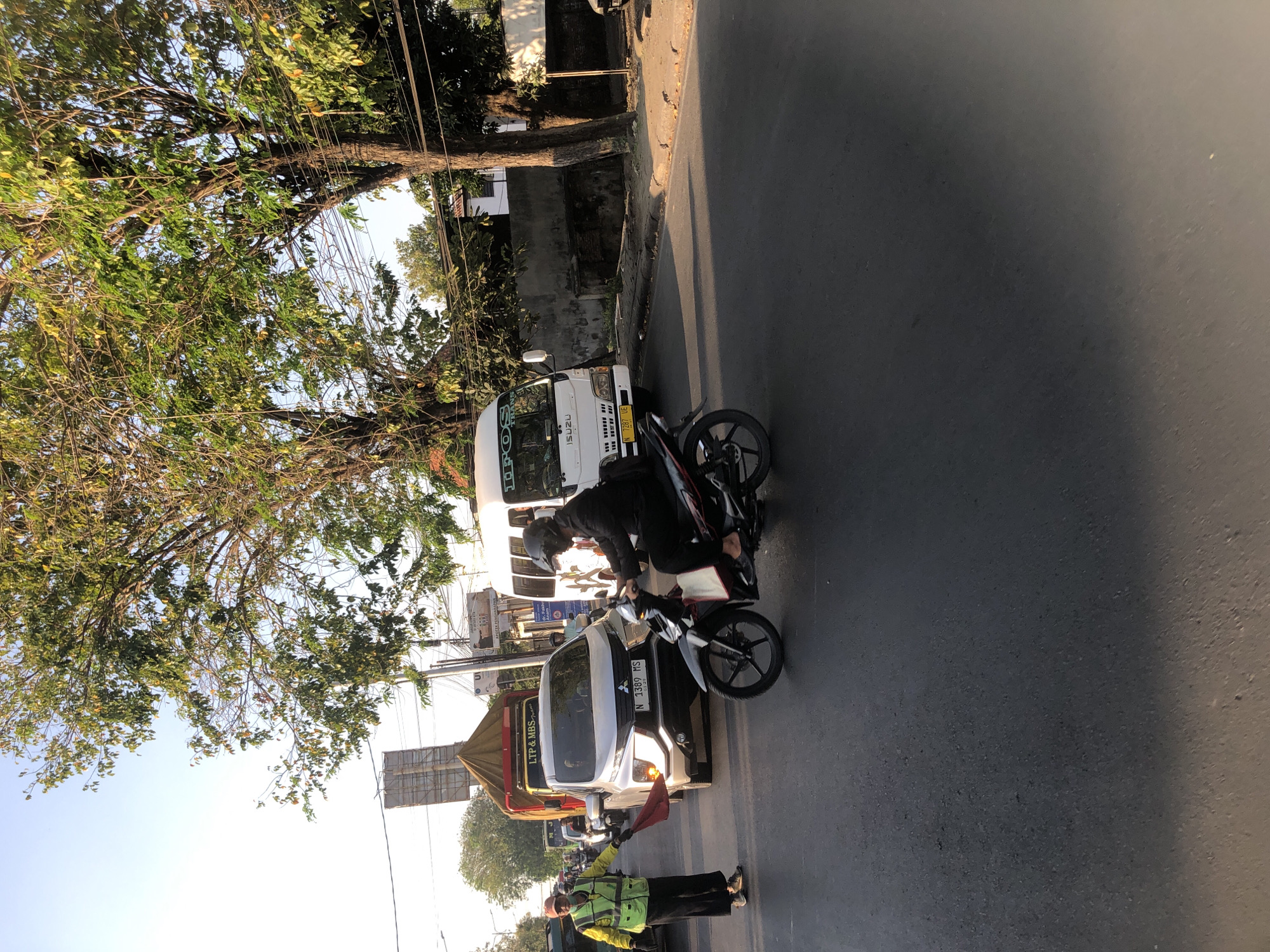 In Kludan, Indonesia, a traffic director in a bright yellow vest and black pants is seen on the left side of the road, holding a red flag. He appears to be directing traffic. In front of him, a silver SUV is stopped, with its headlights on, and a truck with a yellow canopy is behind it. To the right of the SUV, a man wearing a black jacket and helmet is riding a motorcycle. In the background, a white Isuzu bus is visible, along with other vehicles and trees. The scene depicts a street intersection with moderate traffic flow, illuminated by the sun.