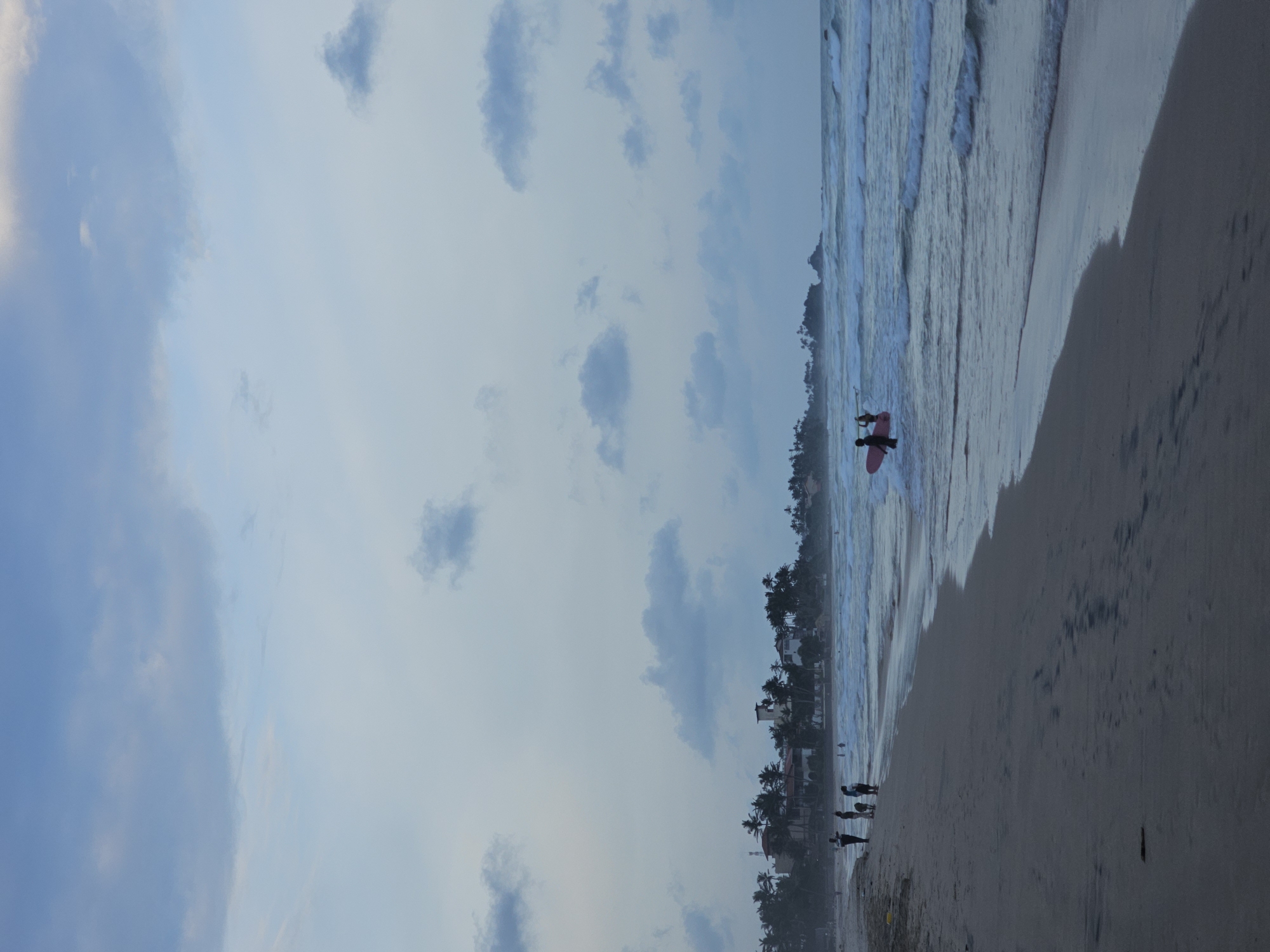 The image shows a beach scene at Kathaluwa, Sri Lanka. The sky is cloudy, and the ocean has gentle waves. A person carrying a red surfboard is wading into the water. Along the shoreline, there are palm trees and buildings. Several people are standing on the sand closer to the edge.