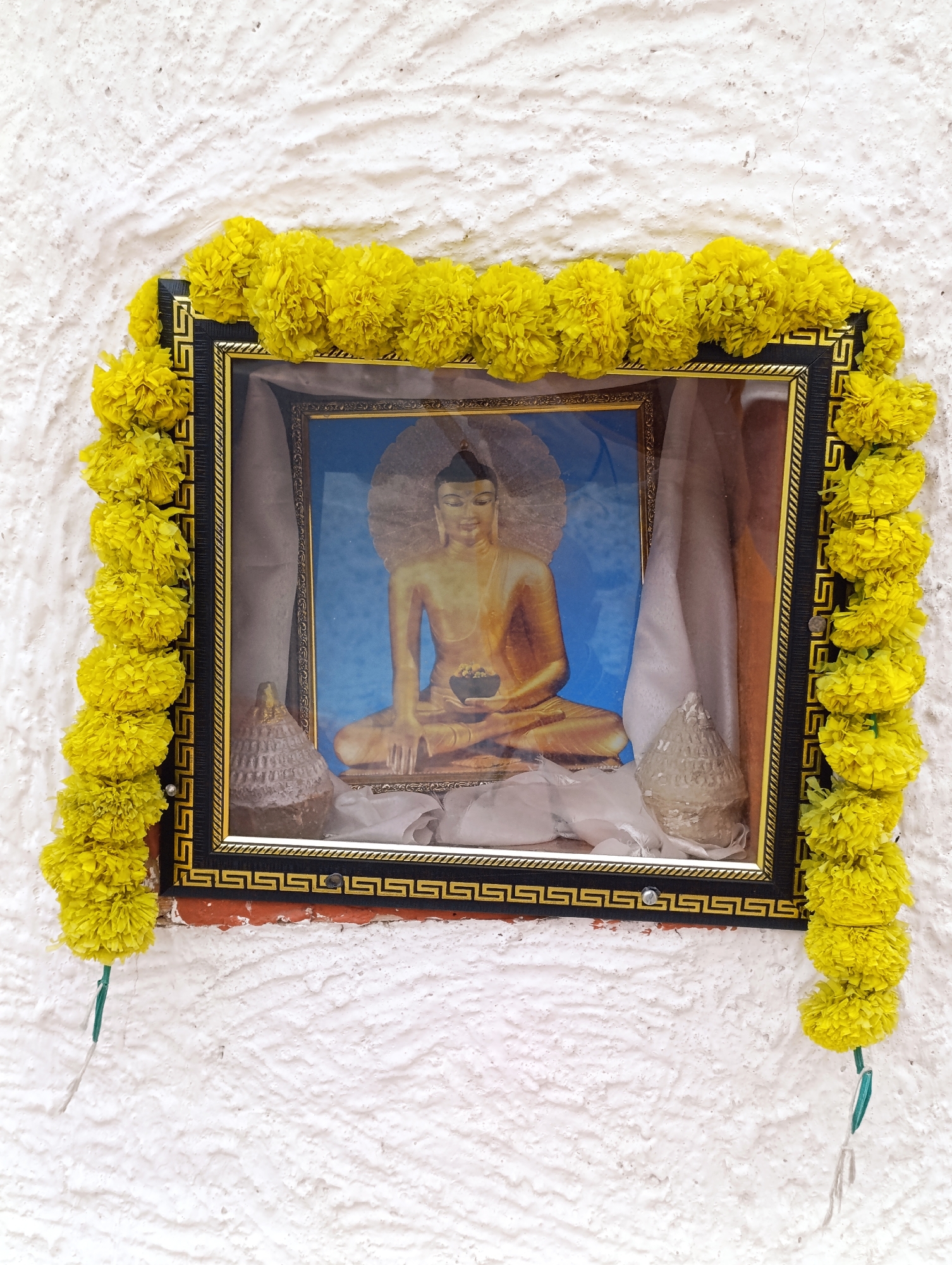 A framed image of a seated Buddha, adorned with a garland of yellow marigolds, is mounted on a rough, white stucco wall in Leh, India. The framed image is rectangular and features a black frame with a gold inner border, inset into a recessed niche. Inside the niche, white fabric drapes behind the image, and two small, cone-shaped objects with a textured, grey surface are placed on either side of the Buddha image. The marigold garland encircles the entire frame, with strands of green string extending downwards from the bottom of the garland. The wall shows signs of texture and some minor imperfections.