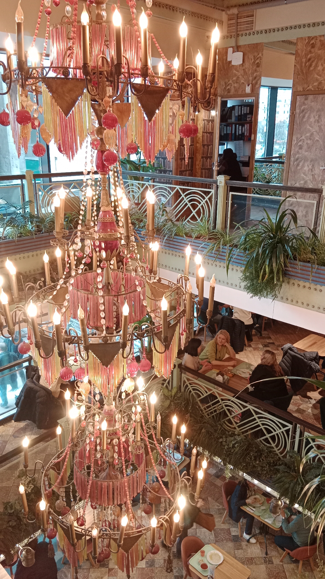 The image shows the interior of a multi-level restaurant in Unknown, Unknown, featuring a large, ornate chandelier as a central focal point. The chandelier is multi-tiered with numerous lights, beads, and decorative tassels. Patrons are seated at tables on different levels of the restaurant. Plants are placed along the railings, adding a touch of greenery to the space. People are visible in different areas, some seated at tables conversing and others passing through the space. The flooring is patterned tile. The overall atmosphere appears upscale and inviting. In the background, a doorway leads to an office or storage area containing shelves of binders or files.