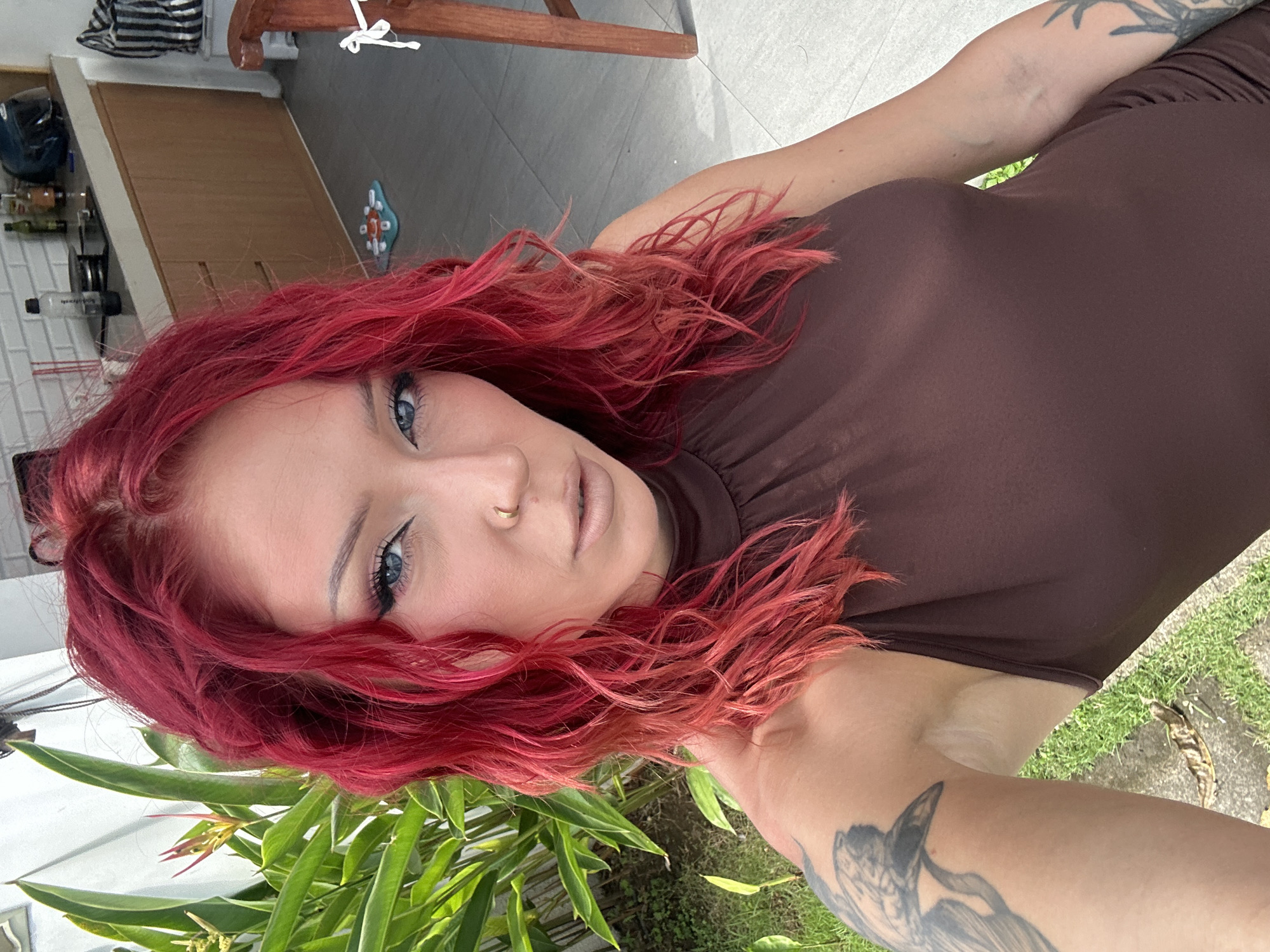 A woman with red hair, blue eyes, and a nose ring is wearing a brown dress. She has tattoos on her arms. She is standing outside among green plants. The location is North Kuta, Indonesia. The background includes a building with tiled flooring.