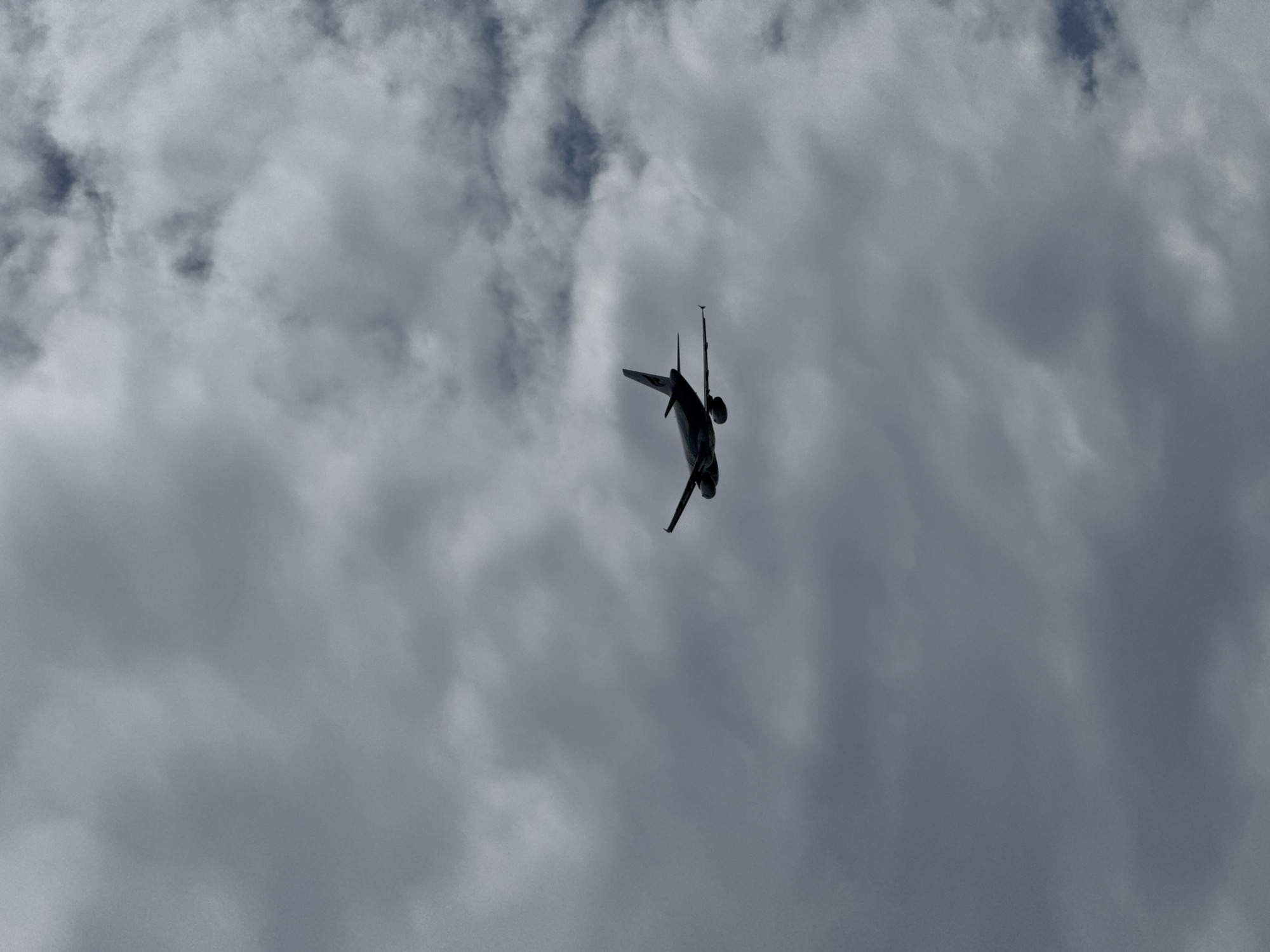A silhouette of an airplane is visible against a cloudy sky, captured from below. The aircraft is angled slightly upwards, with its wings extended horizontally. The clouds are dense and gray, suggesting an overcast or possibly stormy day. The image does not provide any specific landmarks or indications of the city of Ko Samui District, Thailand.