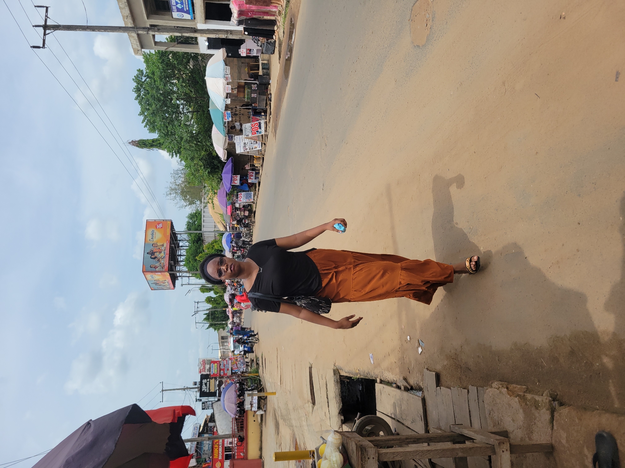 A person stands on a street in Egbagu, Nigeria. The street is paved and there are sidewalks and shops on either side. The shops are covered by umbrellas. The person is wearing a black top and brown pants and glasses. They are holding a blue object in their right hand. There are electrical poles and power lines visible. There is a billboard in the background. There is a mix of cars and motorcycles on the road. There are wooden structures on the ground near the sidewalks.