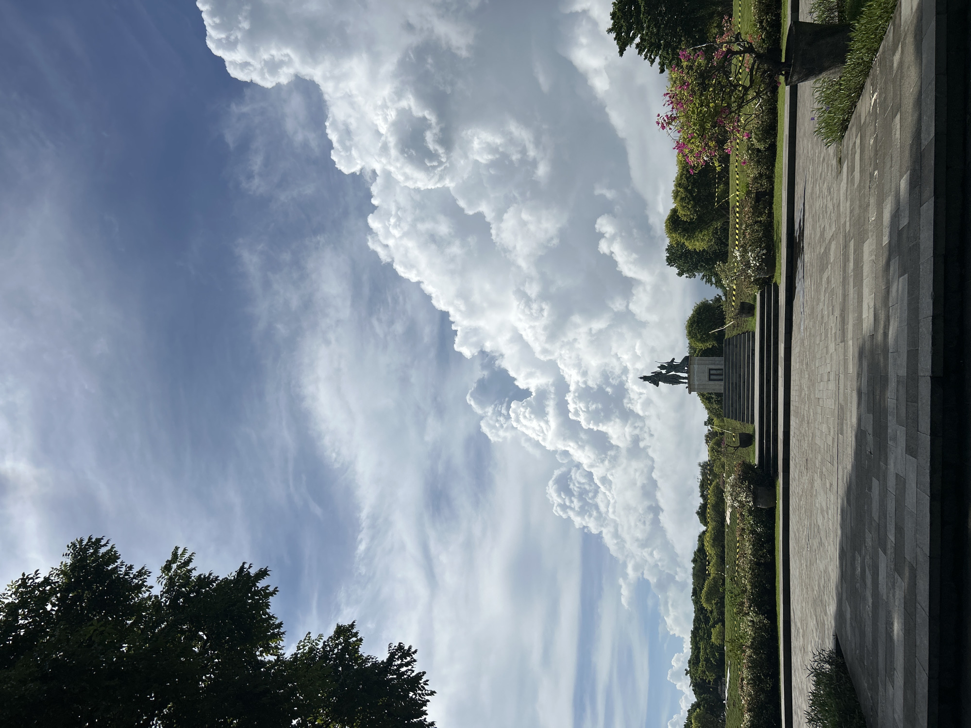 The image displays an expansive outdoor scene under a bright blue sky filled with captivating cloud formations. In the upper right quadrant, a colossal, billowy bank of white cumulus clouds dominates the sky, exhibiting soft, voluminous textures. The upper left and central portions of the sky feature a lighter blue hue with delicate, streaky cirrus clouds.
The lower half of the frame reveals a meticulously maintained landscape. On the right, a broad pathway or plaza constructed from dark gray rectangular pavers extends from the foreground, casting distinct shadows. Parallel to this paved area lies a strip of vibrant green grass and garden beds, adorned with various bushes and plants, some bearing clusters of pink and purple blossoms. Yellow caution tape is visible around some of these planted sections.
Further into the mid-ground on the right, a dark, multi-tiered stone structure or building serves as the base for a prominent bronze-colored statue. The statue, though somewhat distant, appears to portray multiple figures, possibly mounted, indicative of a memorial or monument. The bottom left and far-left edges of the frame are anchored by the dark green foliage of tall trees, providing a natural contrast to the bright sky.
The scene is illuminated by strong sunlight, suggesting it is daytime, either morning or afternoon, under clear weather conditions with the notable cloud patterns. The overall impression is one of a serene and well-kept public park or memorial site, consistent with a location like Kuta Selatan, Indonesia. No discernible text is visible in the image.