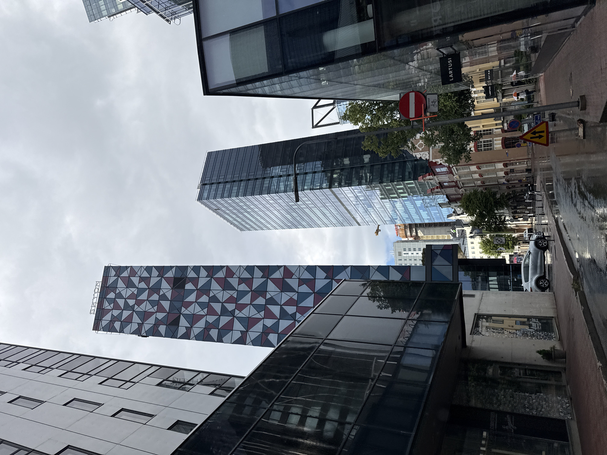 The image shows a street scene in Tallinn, Estonia, with modern skyscrapers and older buildings. In the foreground, a wet street reflects the cloudy sky and surrounding architecture. A silver car is parked on the right side of the street. A dark, modern building with a glass facade occupies the immediate right foreground, with a sign reading "LARTUSI" visible on its exterior. To the left, a sidewalk leads past a building with dark, reflective windows and a concrete base. A tall, slender skyscraper with a patterned facade of red, blue, and grey triangles dominates the midground, identified by the "SKYON" sign at its top. To its right, a dark, glass-covered skyscraper rises. In the distance, other modern buildings and some older, ornate structures can be seen. The overall atmosphere is urban and somewhat somber due to the overcast sky and wet pavement, suggesting recent rain. Two individuals are visible walking in the distance, one holding a red and white umbrella. Traffic signs, including a "no entry" sign with a bicycle symbol and a triangular warning sign with two arrows, are present on poles. Trees are scattered along the street and sidewalk.