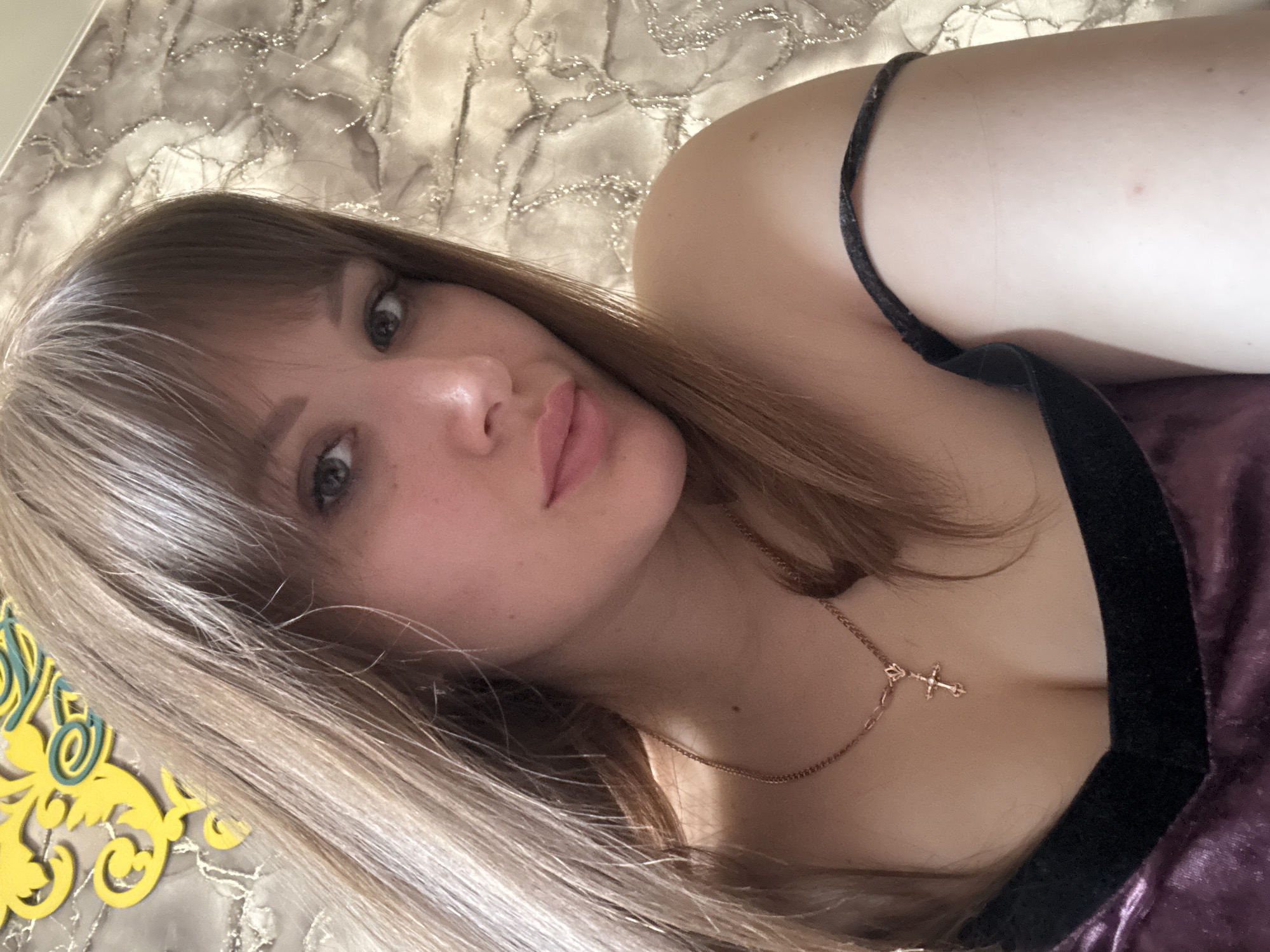 A selfie angled slightly to the left features a woman with light brown hair and bangs. She has fair skin, a pink-toned nose and lips, and green eyes. She wears a dark velvet top with a thin black strap visible on her shoulder. A delicate gold necklace with a cross pendant adorns her neck. The background appears to be a textured surface, possibly a headboard or wall covering with a marble-like pattern. A yellow decorative item with a floral design is visible in the lower-left corner. The location of the city of Chaikovsky, Russia cannot be verified within the context of the image.