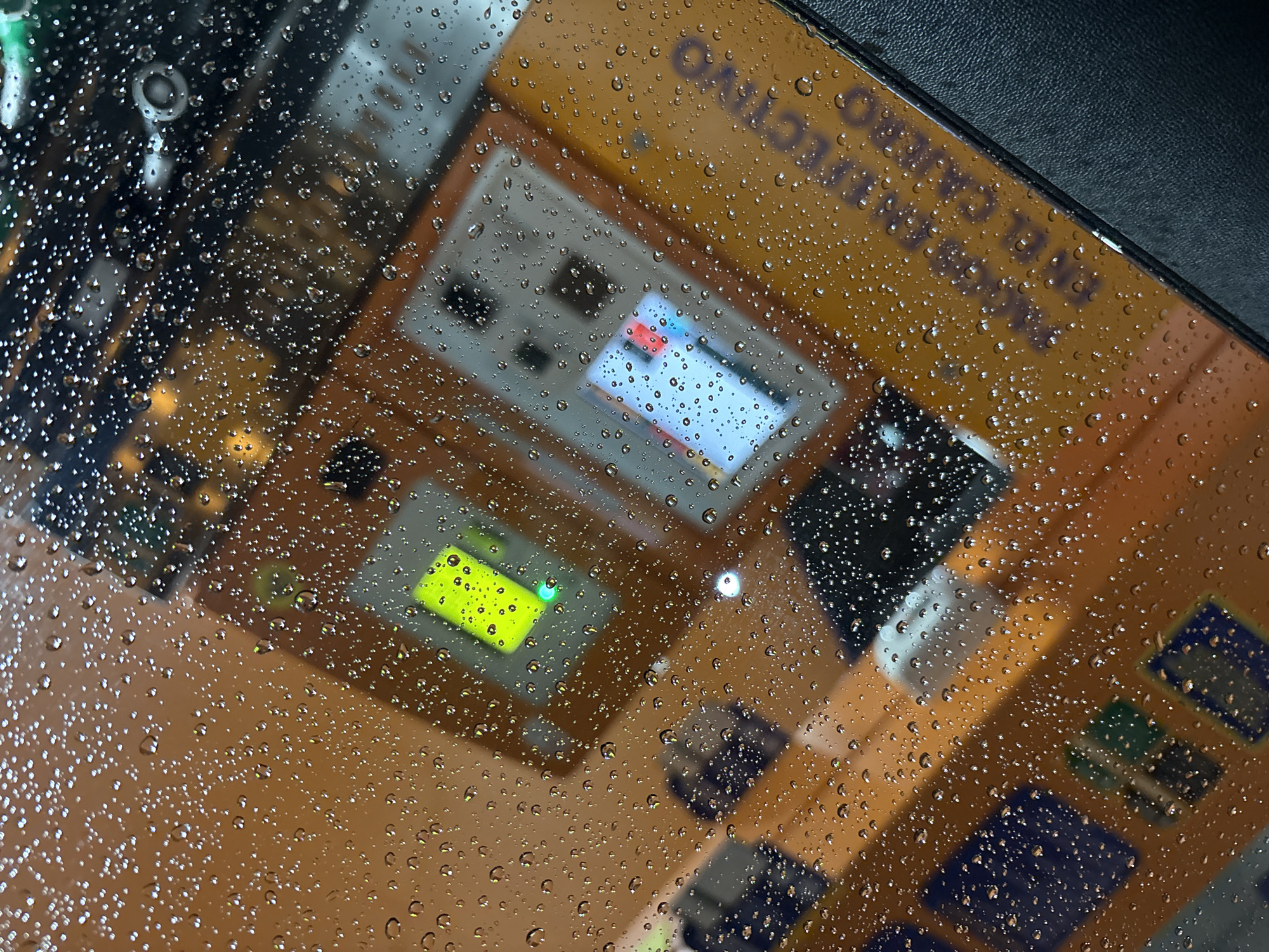 The image shows a rain-covered window reflecting a Caja Rural ATM. The reflection reveals the ATM's interface, which displays various screens and buttons. The environment appears to be a rainy day in Córdoba, Spain, given the text "CÓRDOBA CAJA" visible on the ATM. The ATM's structure is composed of various panels and components. No people are directly visible in the reflection.