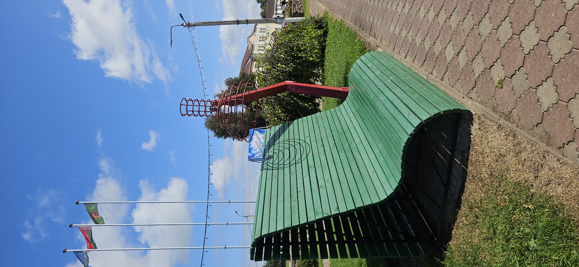 In the city of Liozna, Belarus, under a bright blue sky dotted with white clouds, a distinctive green wooden bench with a curved design is prominently featured. The bench has a textured surface with visible wood grain and is supported by a dark metal frame on its left side. To the right of the bench, a red abstract sculpture resembling a coiled spring or a stylized flame rises from the ground. The sculpture has a light bulb at its center, enclosed by a red wire cage. In the background, a paved walkway made of interlocking reddish-brown tiles extends to the right. Beyond the walkway, there is a patch of grass, some overgrown with dried vegetation on the right side. Further in the background, a building with a light-colored facade and a tiled roof is visible, surrounded by lush green trees and bushes. To the left of the bench, three flagpoles stand tall, each flying a flag: the leftmost flag is blue with a yellow emblem, the middle flag is red and white, and the rightmost flag is green with a yellow emblem. A street lamp with a dark curved arm is situated on the right side of the image, adding to the urban park-like setting. A string of small lights is suspended between the flagpoles and the street lamp. A blue and white banner with text and an image of clouds is also visible behind the bench and the red sculpture. The overall scene depicts an outdoor public space on a sunny day.