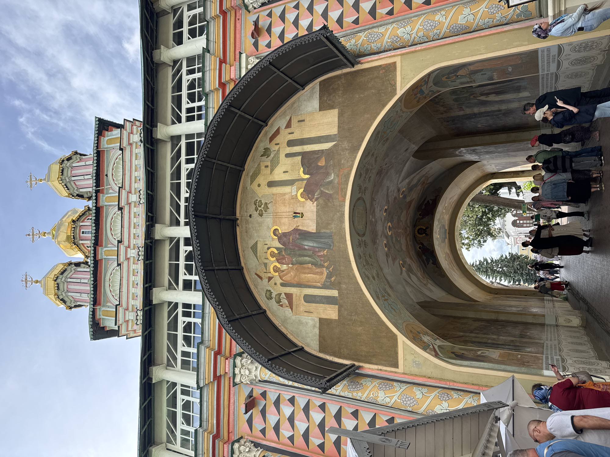 The image depicts the Trinity Lavra of St. Sergius in Sergiev Posad, Russia, specifically showing a section of the complex with religious iconography and architectural details. In the foreground, an arched entryway is visible, adorned with frescoes depicting religious scenes and figures with halos. The walls of the entryway are decorated with geometric patterns and vine motifs in vibrant colors. Above the entryway, a building with a colorful facade, featuring striped patterns and white window frames, is topped by golden domes with crosses, indicating its ecclesiastical function. People are seen walking through the arched passage and congregating in the area, some appearing to be tourists. The lighting suggests it is daytime, with a partly cloudy sky.
