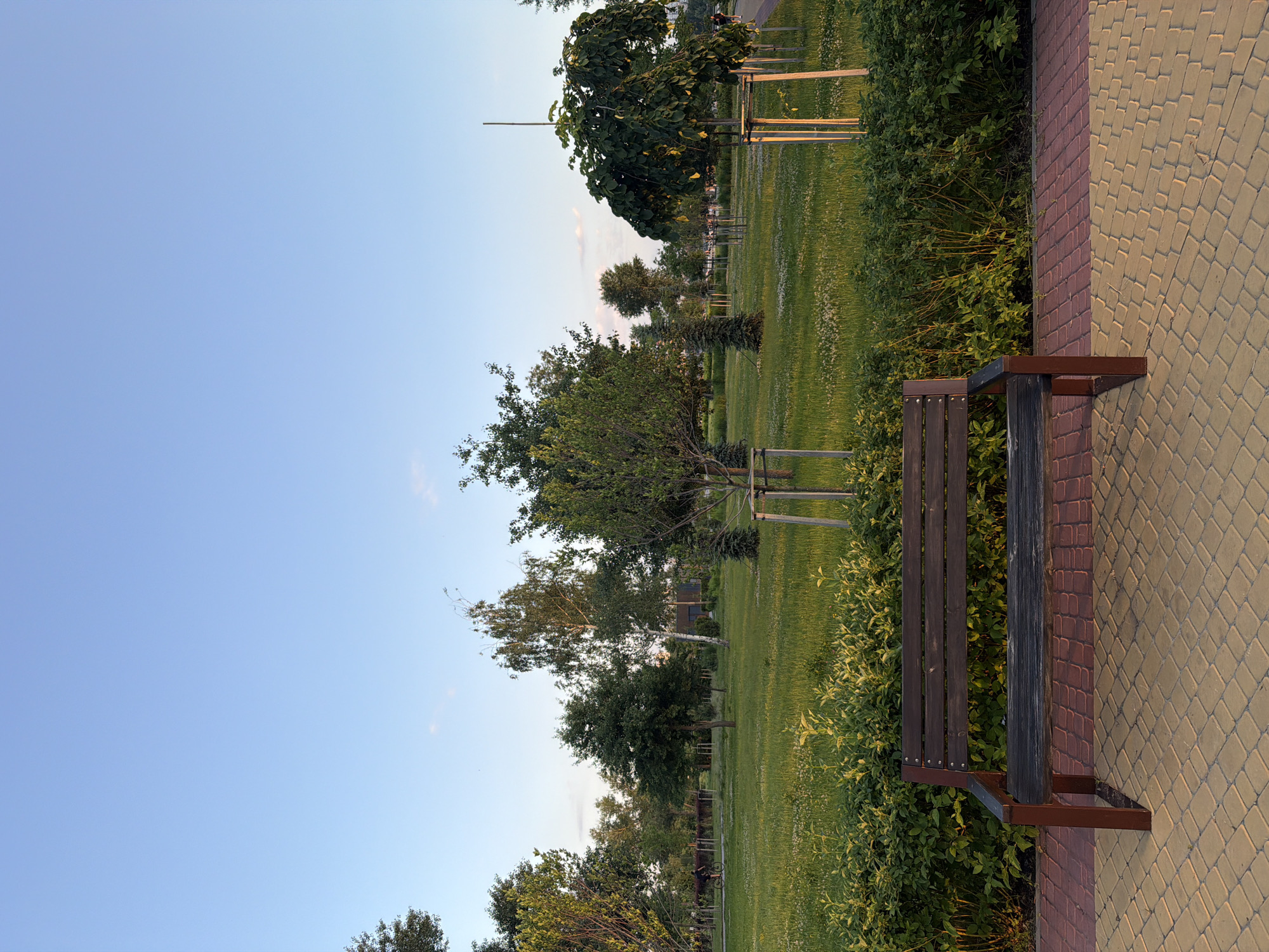 The image shows a park with a paved walkway in the foreground. A wooden bench is positioned on the walkway, facing a grassy area with scattered trees and bushes. The sky is clear and blue, suggesting it is daytime. In the distance, a person is riding a bicycle on a path. This scene is located in Kyiv, Ukraine.