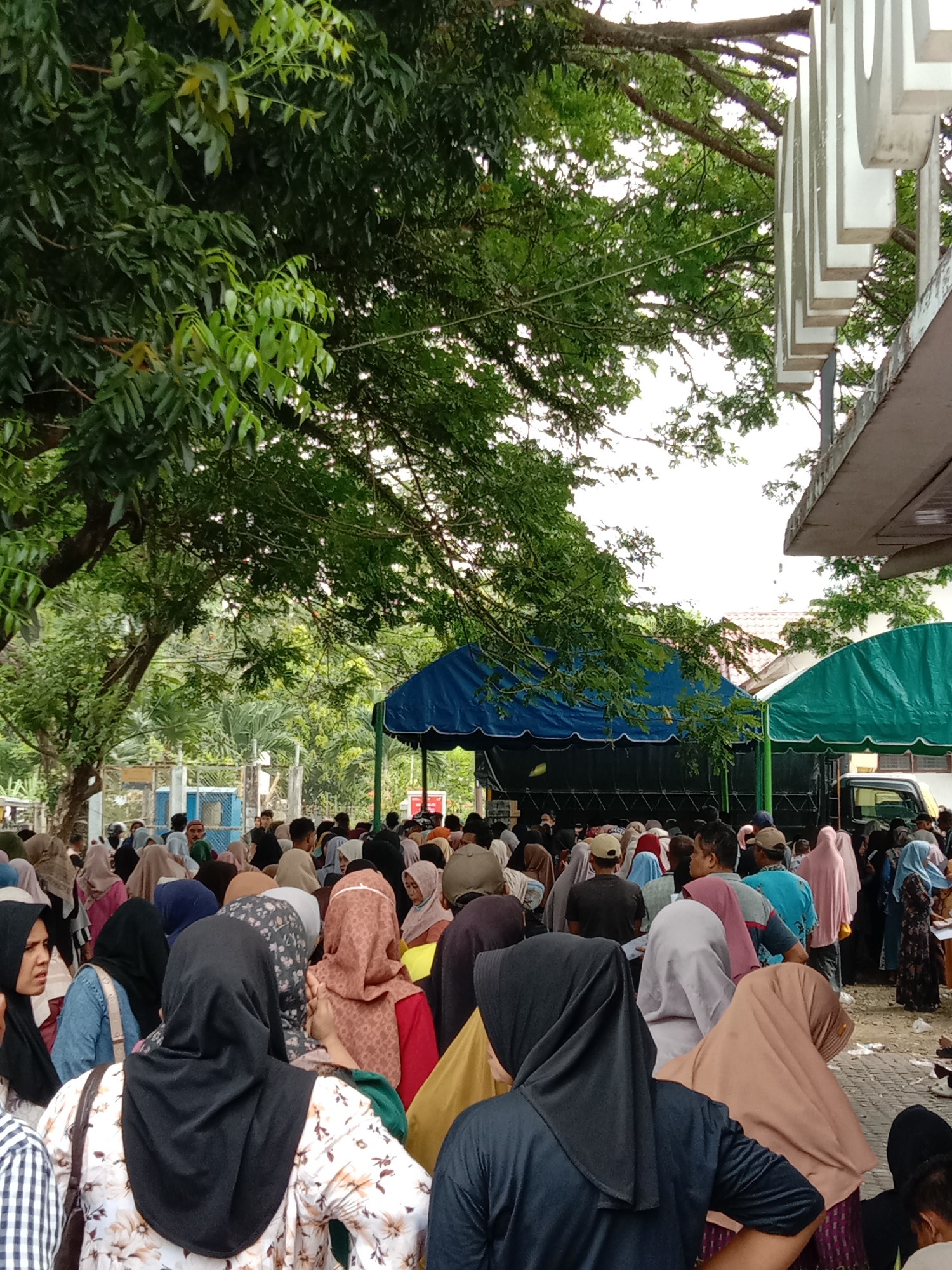 A crowd of people, many wearing hijabs, are gathered outdoors near what appears to be a distribution point under blue and green canopies. A truck is parked adjacent to the green canopy. Large trees provide shade. The location is identified as Lhokseumawe, Indonesia.