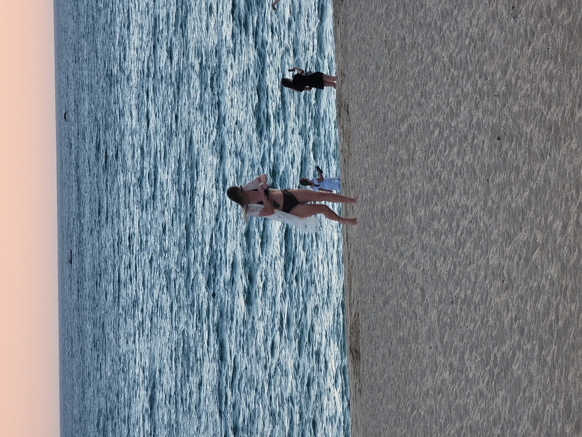 A vertical, eye-level, medium shot captures a beach scene in Quarteira, Portugal, with the calm, choppy ocean stretching to the horizon under a dusky sky. In the foreground, a woman in a black bikini and a sheer white cover-up walks left to right on the sandy shore, holding a phone. To her right, slightly further out, a woman in a black dress stands facing right and holds a phone, possibly taking a photo. In the mid-ground, a person in a light blue garment stands near the water's edge. Two small, dark objects, possibly boats, are visible on the distant water.
