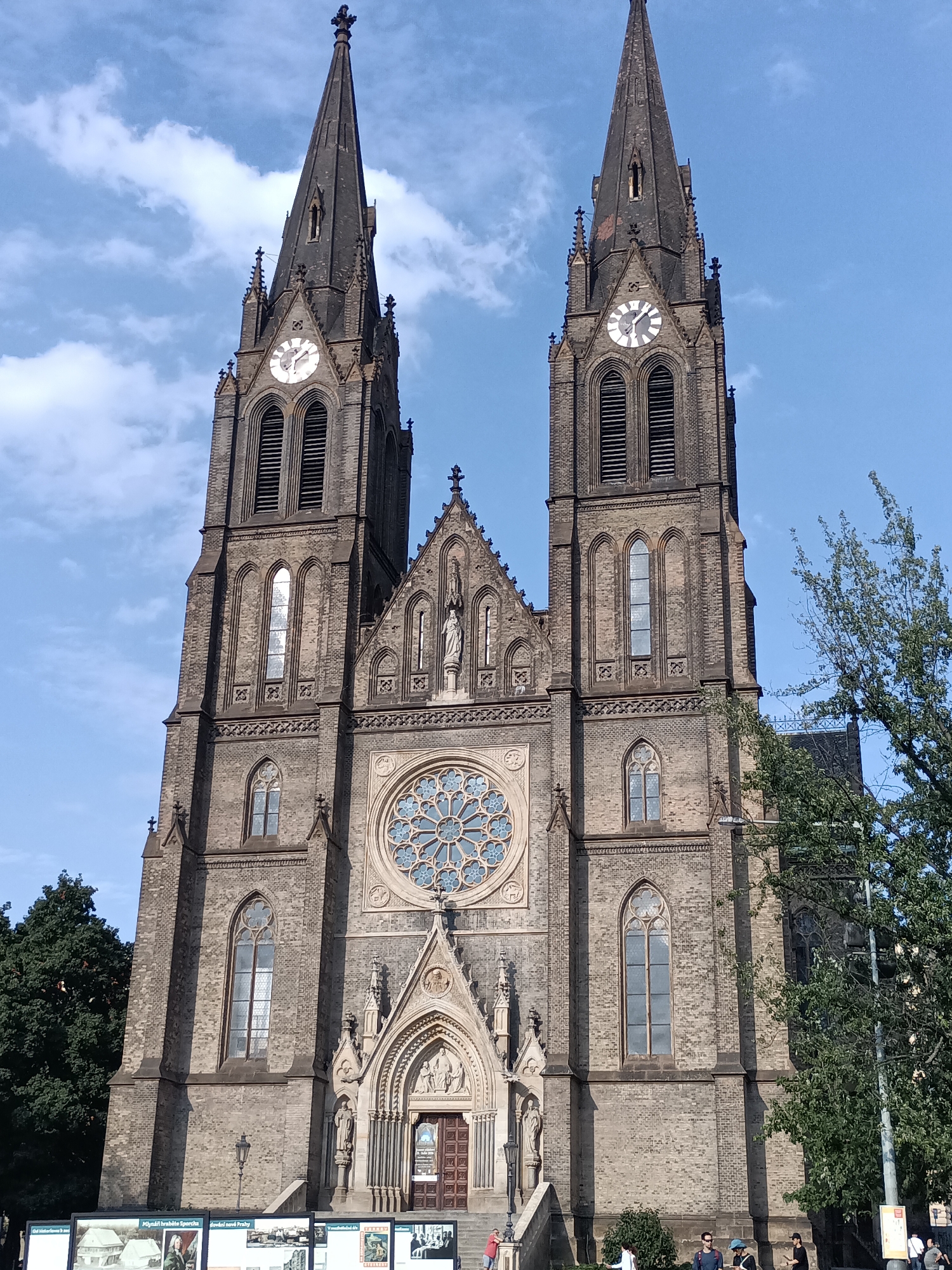 The image shows the facade of the Basilica of St. Peter and St. Paul in Vyšehrad, Prague, Czechia. The basilica is a Gothic Revival Roman Catholic minor basilica. The main facade features two prominent spires, each with a clock face and arched windows. A large rose window is centrally located above the main entrance, which is adorned with sculptures. The building is constructed of brick and has several smaller Gothic-style windows. A statue of a religious figure is positioned above the rose window. In front of the basilica, there are informational display boards. Several people are visible in the foreground, some standing and looking at the displays, others walking. Trees are present on the right side of the image, partially obscuring the view of the basilica. The sky is blue with scattered clouds.