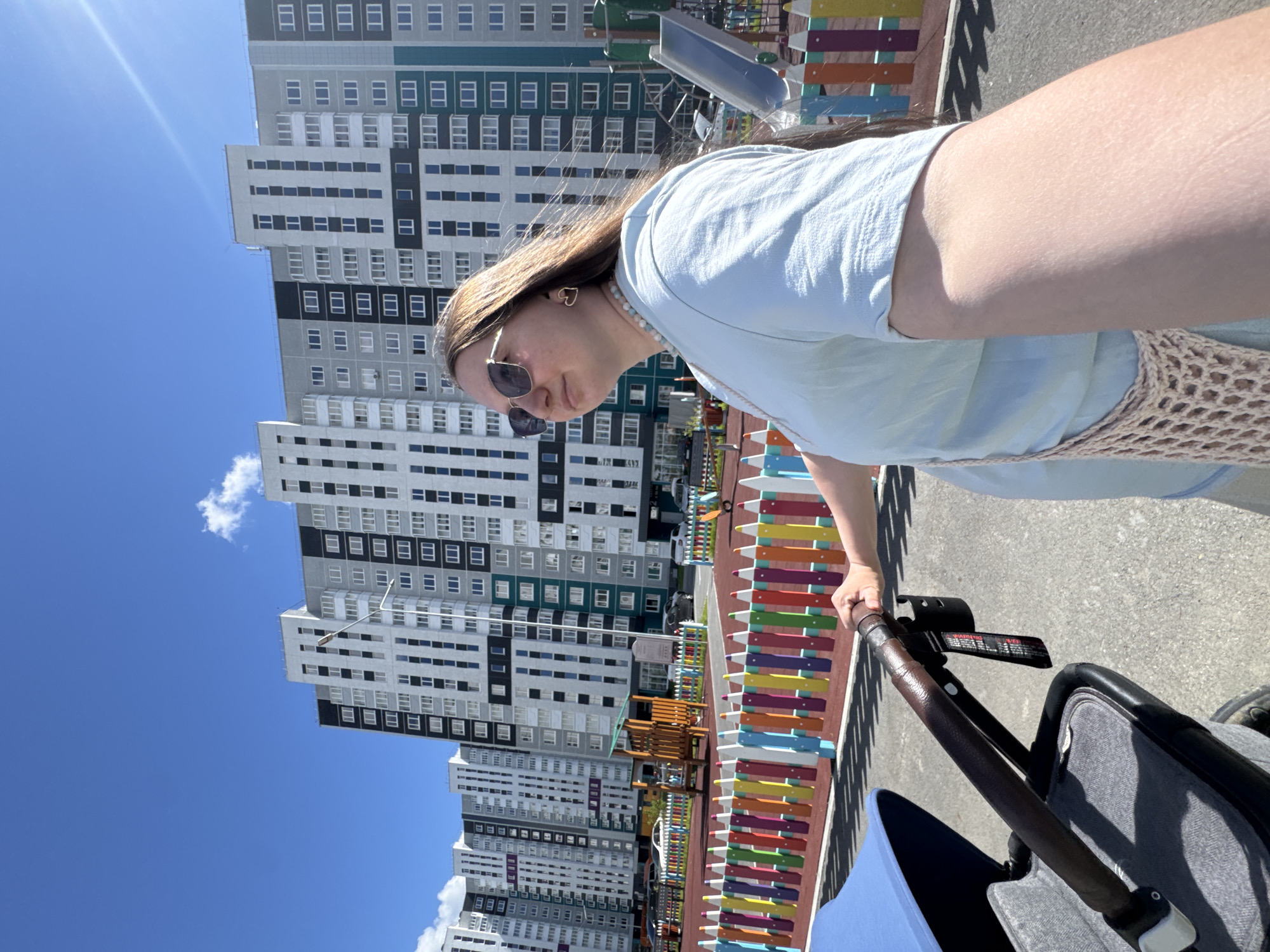 In this photograph, a young woman is captured from a slightly elevated perspective, holding the handle of a stroller. She is wearing a light blue t-shirt and sunglasses, and her hair is long and brown. A beaded necklace adorns her neck, and she has heart-shaped earrings. The background features a modern residential complex with high-rise buildings. To the right of the woman and stroller, there is a colorful fence made of wooden planks in various hues like red, orange, yellow, green, blue, and purple. A playground with wooden structures and a blue slide is also visible behind the fence. The scene is illuminated by bright sunlight, indicating it's daytime. The sky is a clear, vibrant blue with a few wispy white clouds. The ground is paved with asphalt. The overall impression is a pleasant outdoor scene, possibly in a park or residential courtyard. The woman's expression, though partially obscured by sunglasses, suggests a relaxed or content mood. The photograph is taken outdoors in a setting that appears to be a developed urban or suburban area with a focus on residential living and recreational spaces. There is a visible warning label on the stroller handle, but the text is too small to be legible. The location is identified as Patrushiva, Russia.