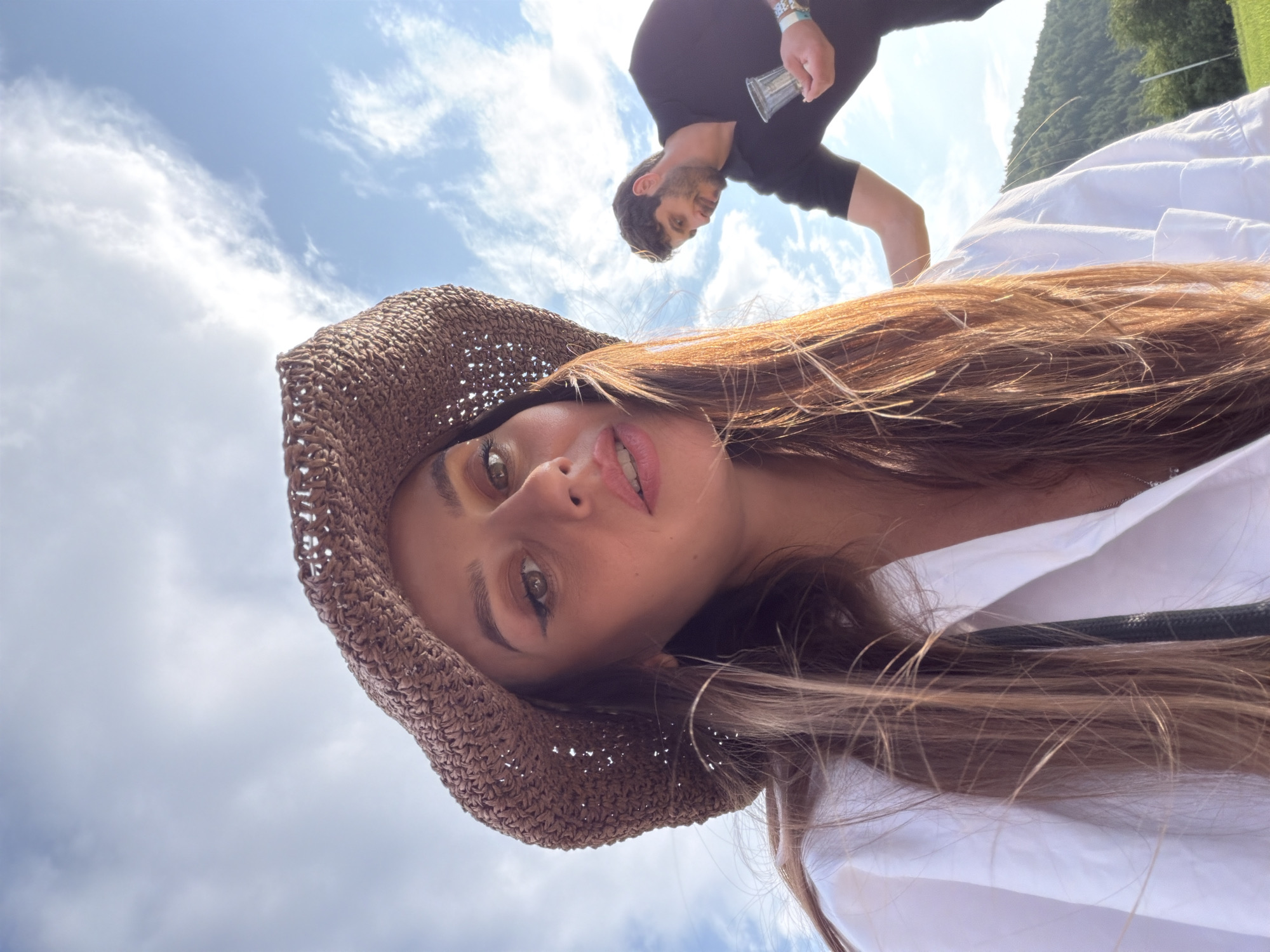 A woman wearing a straw hat and a white shirt is in the foreground of the image, looking towards the camera. Her hair is long and brown, and she has makeup on. In the background, a man in a black shirt is visible, holding a clear plastic cup. The scene appears to be outdoors, with a blue sky and white clouds above. There are trees and a hill in the distance, suggesting a natural or park-like setting. The location is identified as Drokkino, Russia.