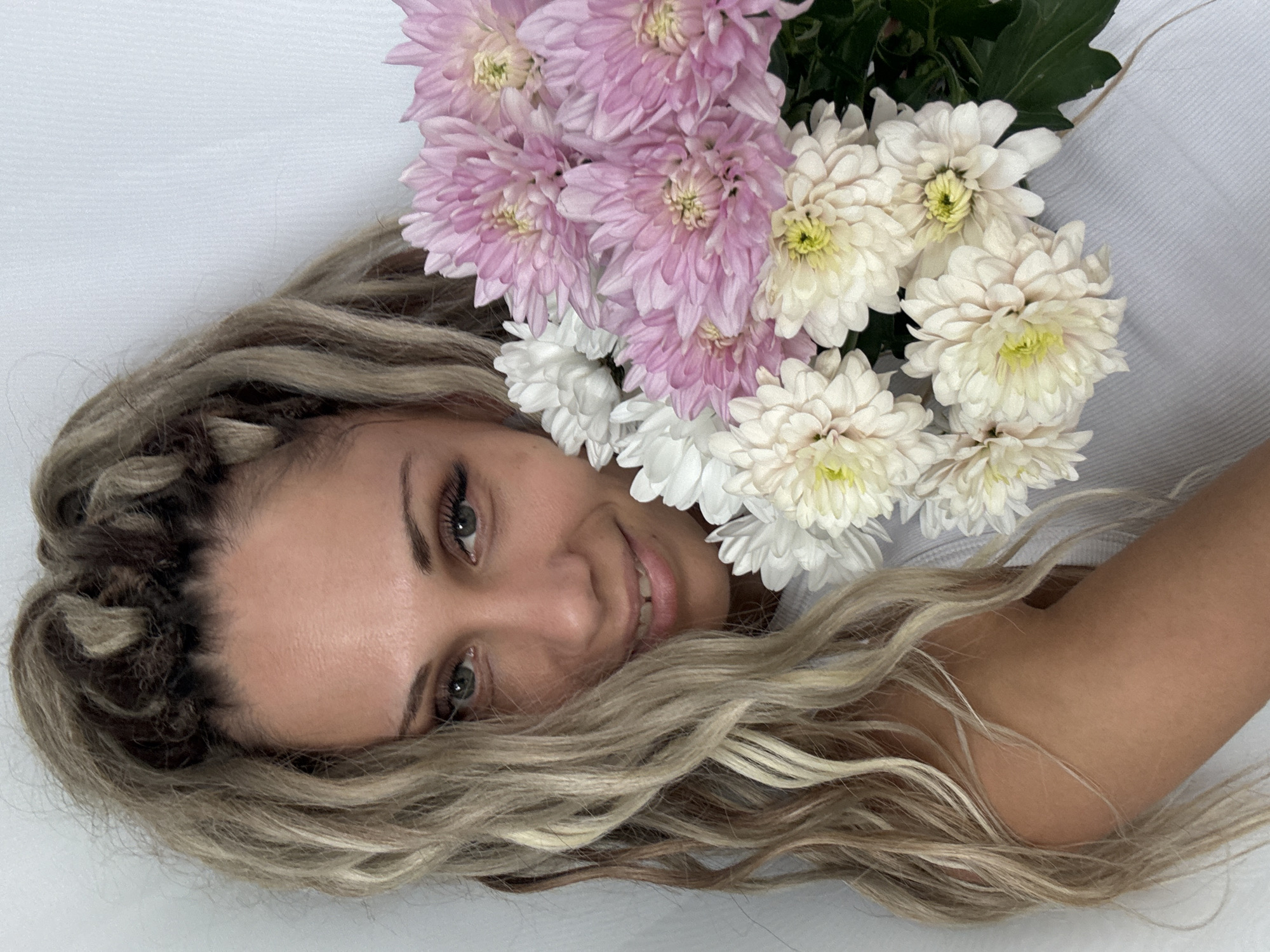 A medium shot shows a woman with blonde dreadlocks, fair skin, and green eyes holding a bouquet of pink and white chrysanthemums. She is smiling and looking towards the camera. The background is a plain white wall. The image does not provide any information about the location in the city of Rusanovo, Russia.