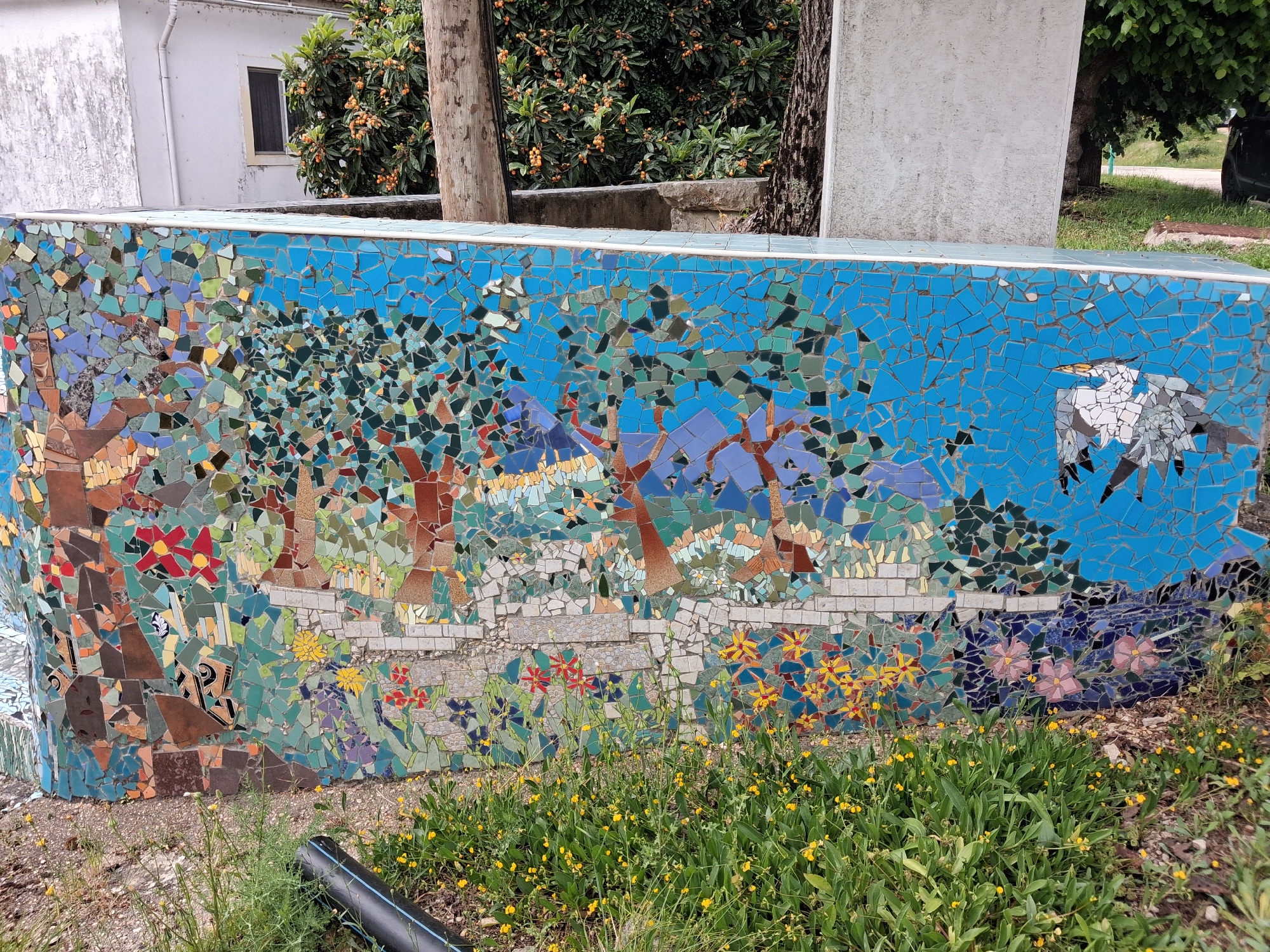 A mosaic wall in Soure, Portugal, depicts a nature scene. The mosaic art includes trees, flowers, mountains, and a bird on a background of blue sky. A retaining wall is behind the mosaic, flanked by a tree and a building. Yellow wildflowers grow along the base of the mosaic. A black pipe lies on the ground beside the wildflowers.