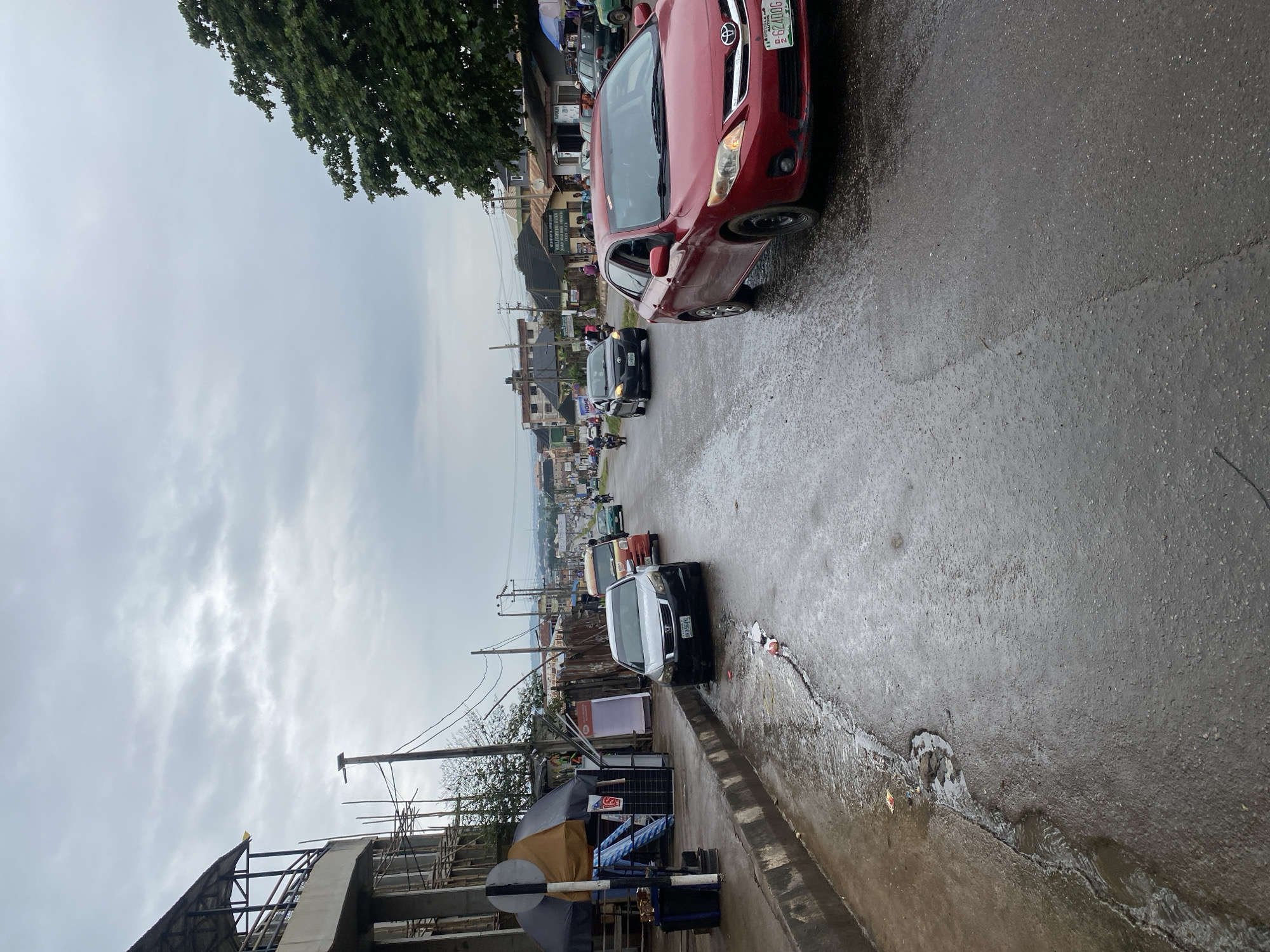 The image depicts a street scene in Abeokuta, Nigeria, under an overcast sky. A wet, asphalt road runs through the center of the frame, with a concrete sidewalk to the left. Several vehicles are present, including a red Toyota Corolla in the foreground, a silver sedan, and an orange mini-bus. A variety of buildings and structures line the street, some appearing to be shops or market stalls. Construction scaffolding is visible on one building on the left. The road surface shows signs of wear and tear, with a crack and a puddle of water. The overall atmosphere suggests a cloudy or rainy day.