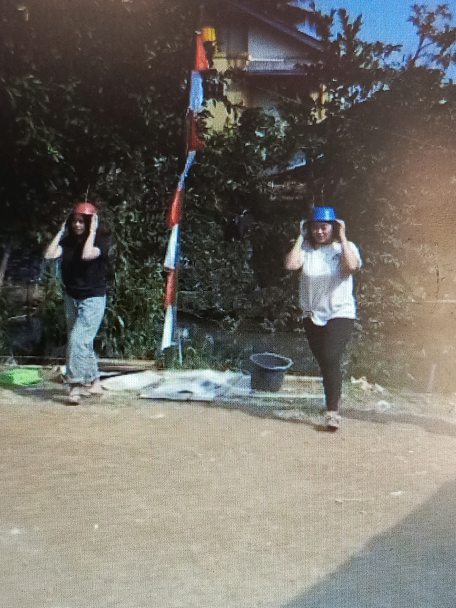 Two individuals are observed in an outdoor setting, with dense foliage in the background and a building visible through the trees. A flagpole with red, white, and blue banners stands prominently between them. The person on the left, wearing a black t-shirt and light-colored pants, is holding a red hat on their head with both hands. The person on the right, dressed in a white t-shirt and dark pants, is holding a blue hat on their head with both hands. Both individuals appear to be standing on a textured ground surface, possibly dirt or gravel. There is a dark bucket visible near the person on the right, and various objects, including what appear to be tarps or mats, are scattered on the ground in the middle ground. The scene suggests an activity or event taking place outdoors. The location is identified as Tik Jeniak, Indonesia.